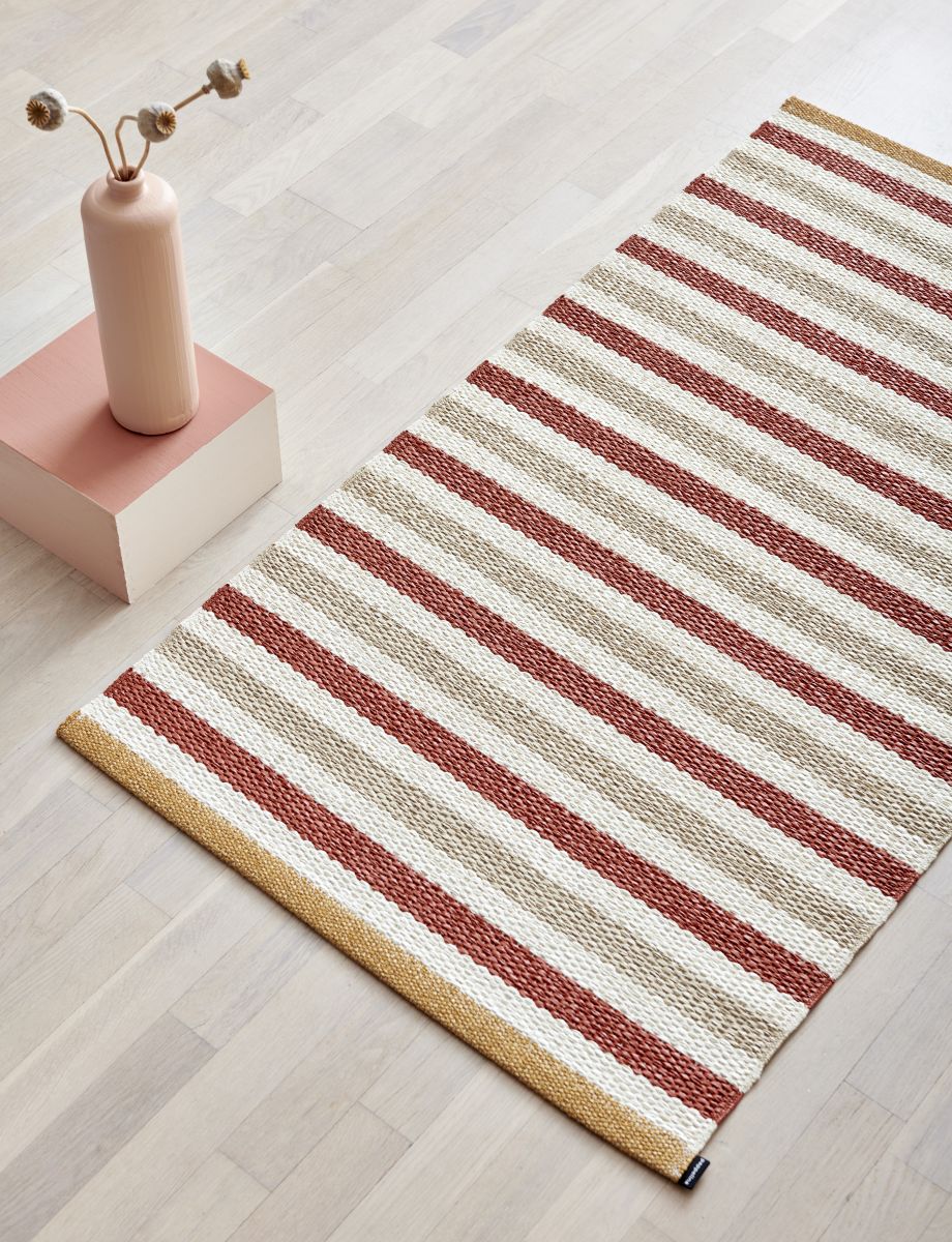 Pappelina Teppich Teo mit Streifen in Beige und Rotbraun auf hellem Holzboden.