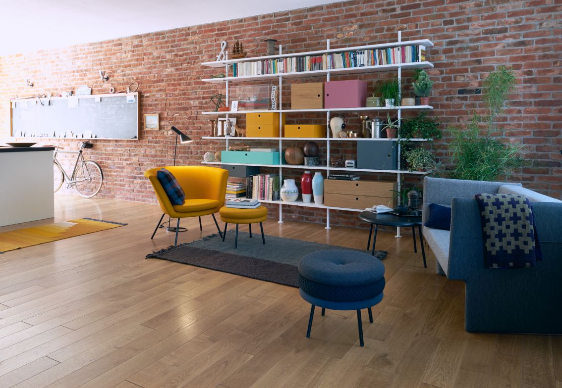 Wohnzimmer mit gelbem Sessel, blauem Hocker, grauem Sofa und weißem Bücherregal vor einer Backsteinwand.