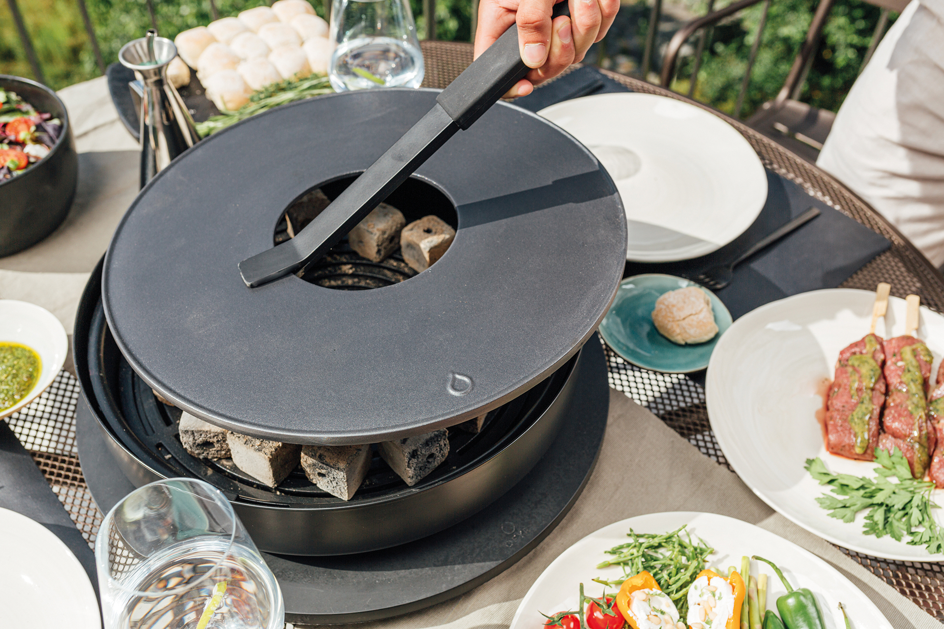 Tabl´O Tischgrill von Ofyr mit Grillzange und Holzkohle, umgeben von Speisen auf dem Tisch.