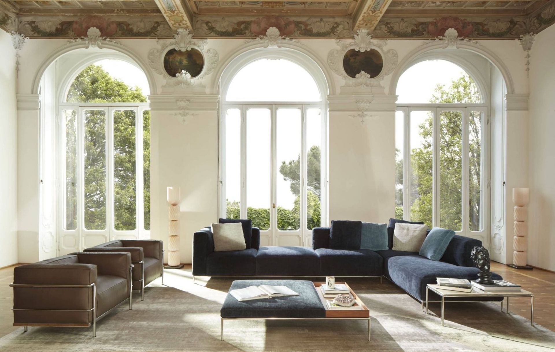 Wohnzimmer mit blauer Eckcouch, braunen Sesseln und Couchtischen vor großen Fenstern. Moderne Möbel.