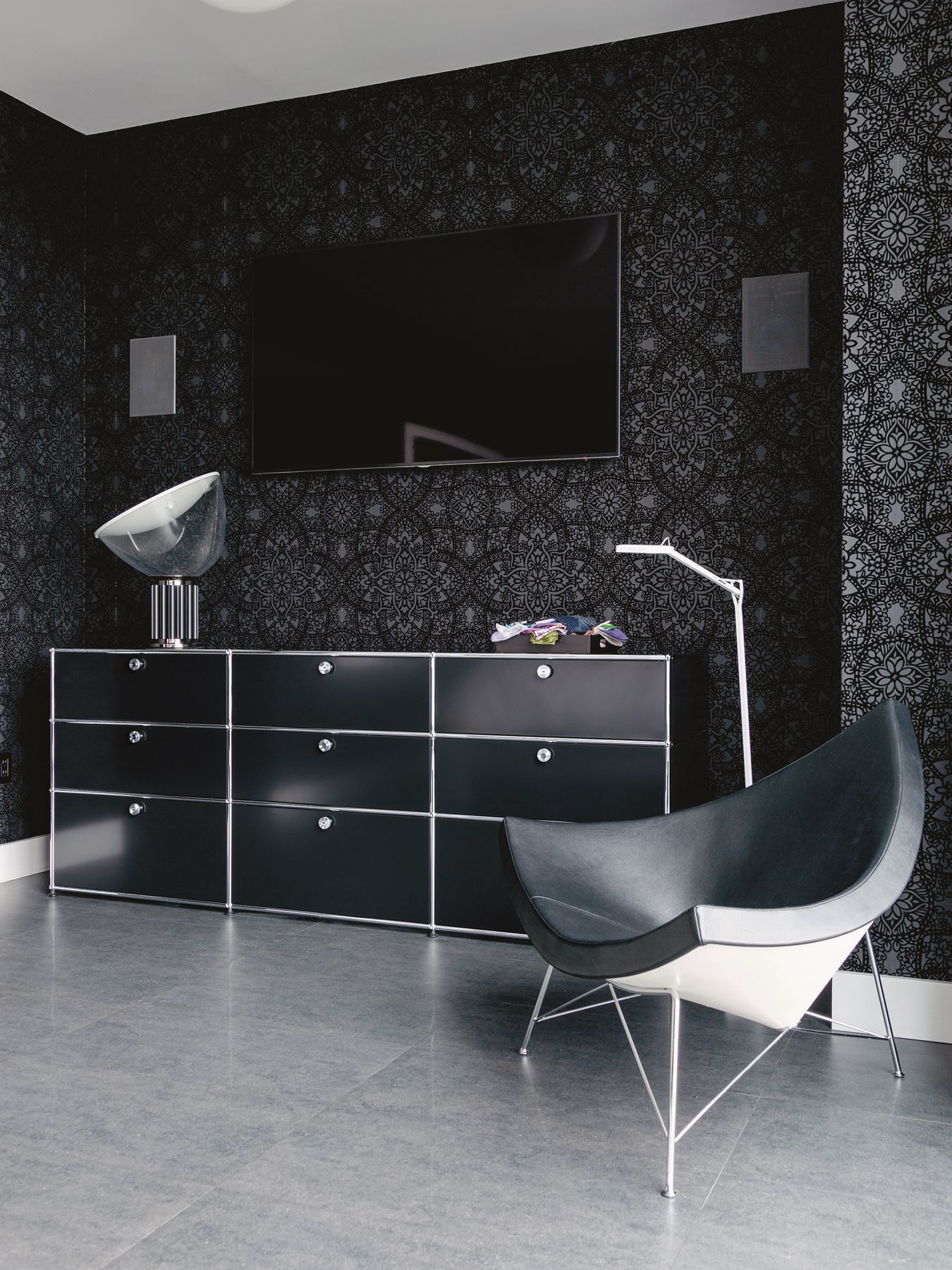Schwarzes Sideboard mit Schubladen, Design Lampe und Ledersessel vor gemusterter Wand im modernen Wohnzimmer.