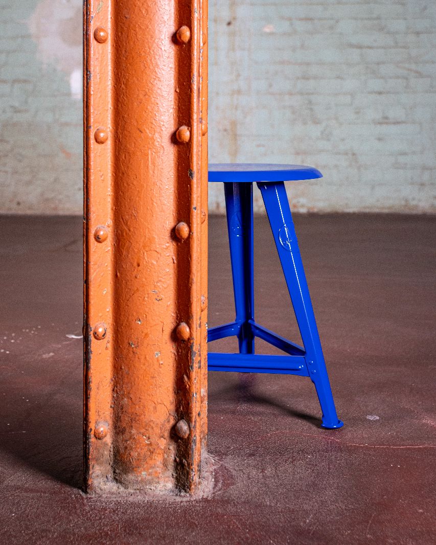 Blauer Rowac Schemel IS 48 cm Hocker Blueprint Edition neben orangefarbenem Pfeiler in Industriehalle.