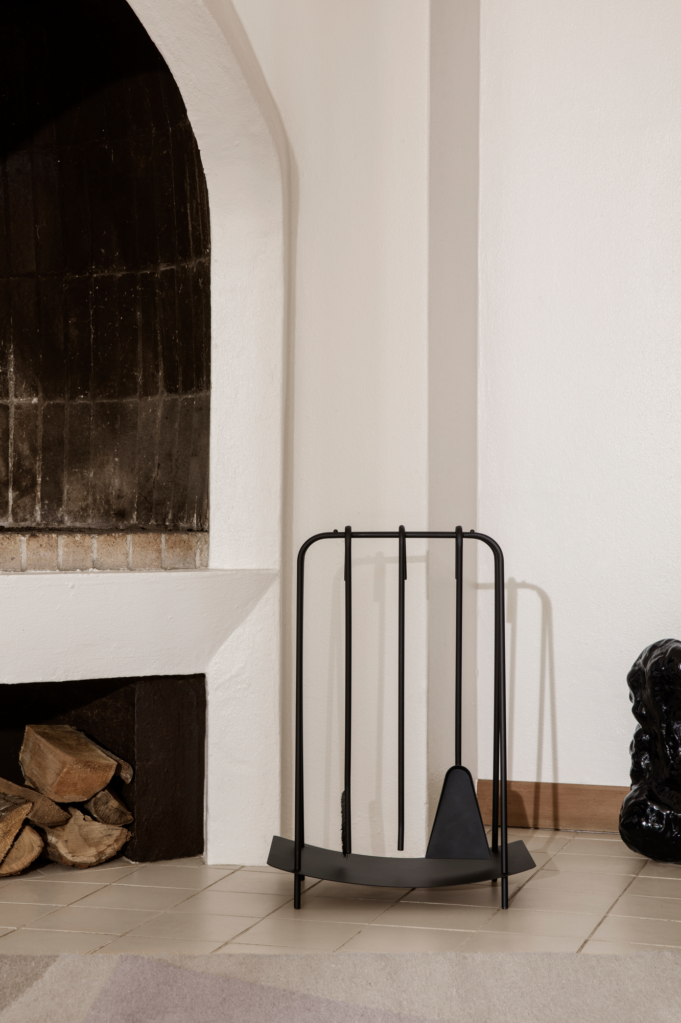 Schwarzes Kaminbesteck von Ferm Living neben einem Kamin mit Holzscheiten. Kaminzubehör für gemütliche Abende.