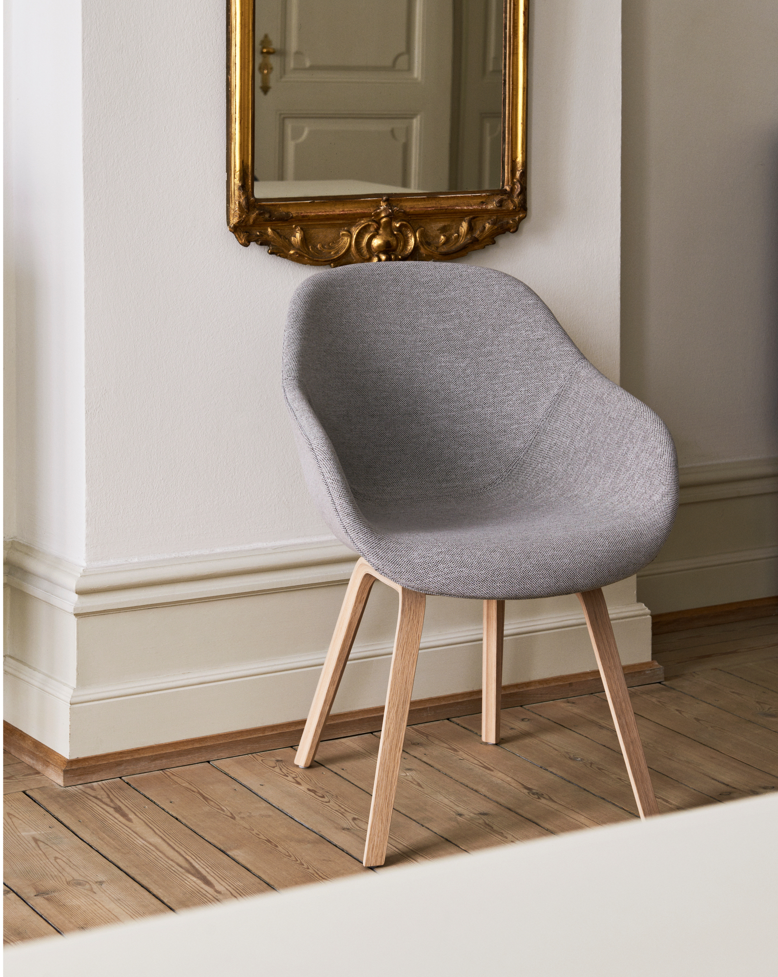 Grauer About a Chair AAC123 Stuhl mit Holzbeinen vor einer Wand mit goldenem Spiegel.