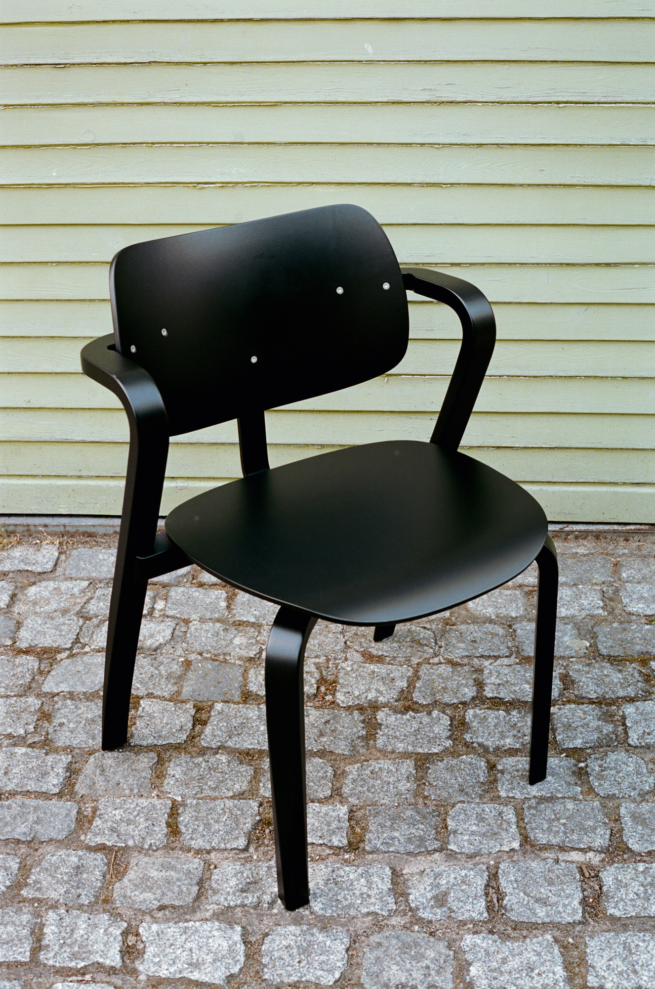 Schwarzer Aslak Chair Stuhl von Artek mit Armlehnen auf Pflastersteinen vor einer Holzwand.