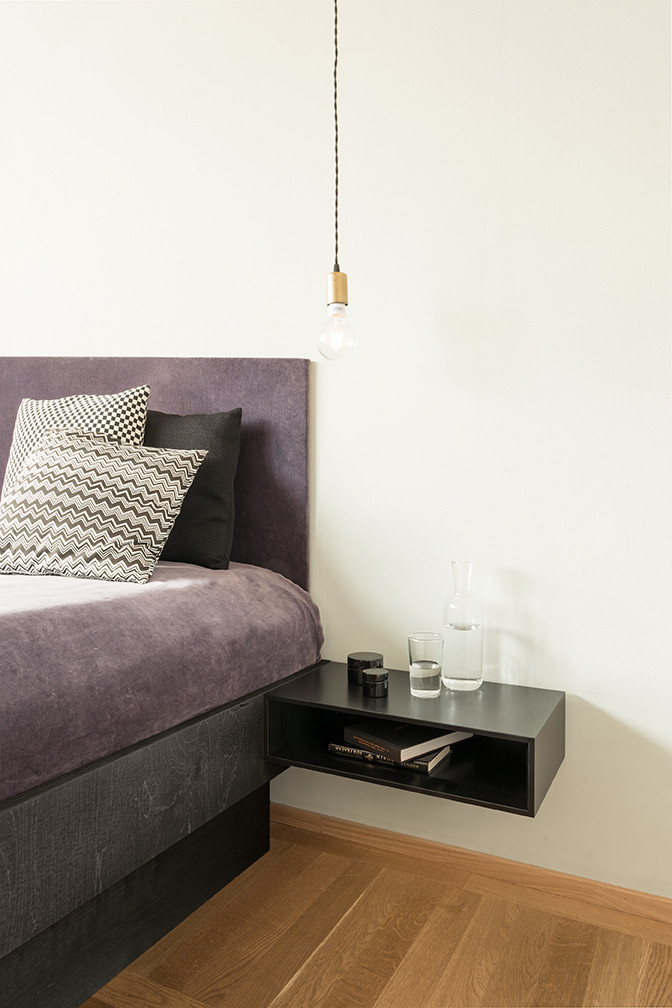 Schlafzimmer mit lila Bett, Nachttisch in Schwarz und hängender Glühbirne für moderne Schlafzimmereinrichtung.