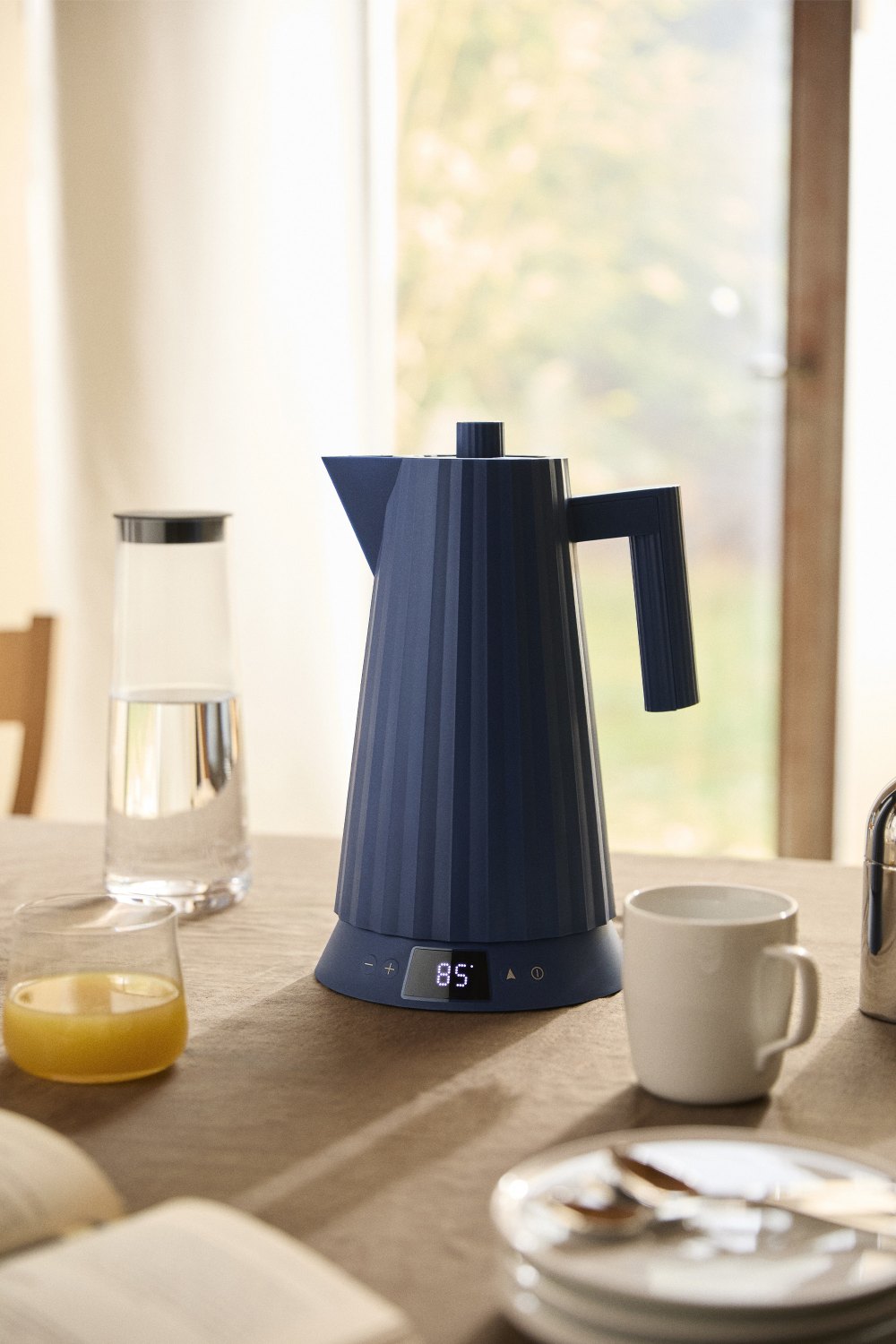 Blauer Alessi Plissé Wasserkocher mit Temperaturanzeige auf einem Tisch mit Frühstücksutensilien.