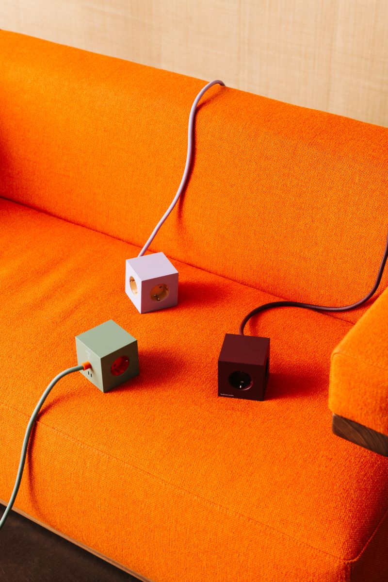 Drei Avolt Square 1 Steckdosenwürfel in Lila, Grün und Rot auf einem orangefarbenen Sofa.