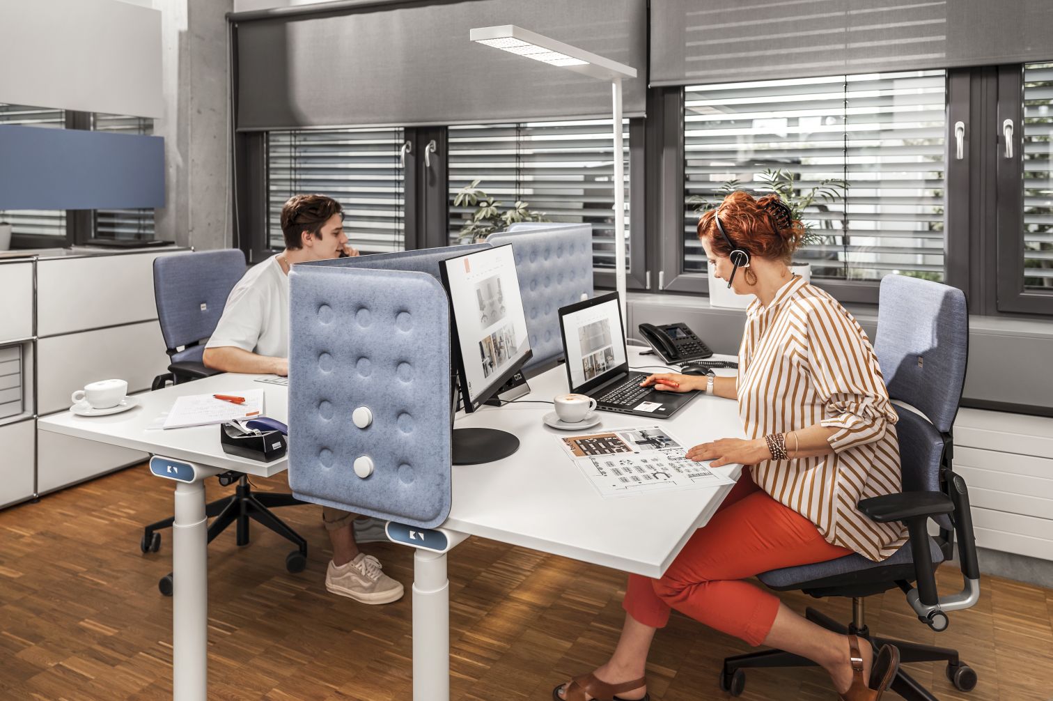 Büro mit höhenverstellbarem Schreibtisch, Akustikpaneelen, ergonomischen Bürostühlen und LED-Schreibtischlampe für modernes Arbeiten.