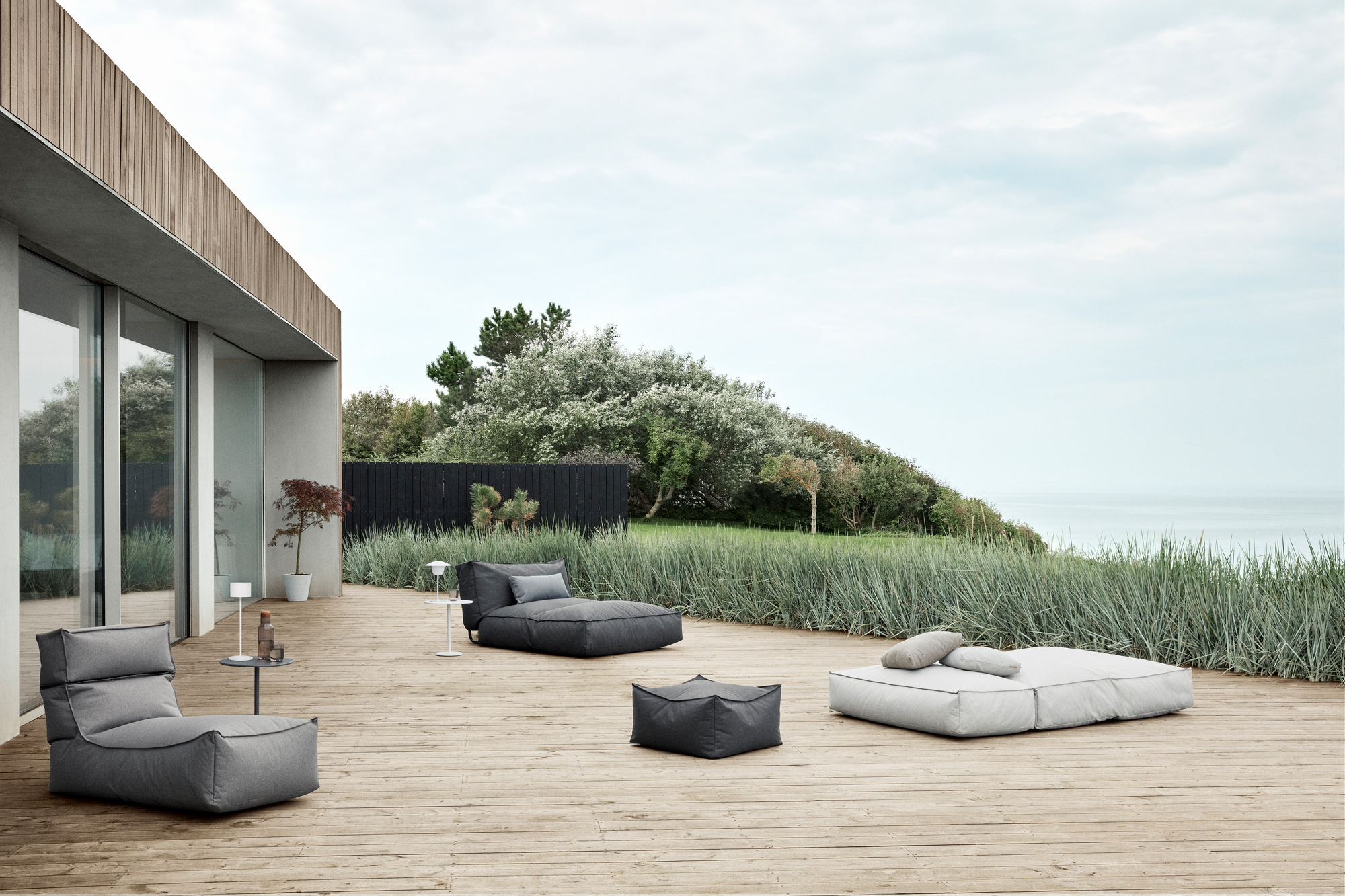 Drei Blomus Stay Outdoor Kissen in Grau und Weiß auf einer Holzterrasse mit Meerblick.