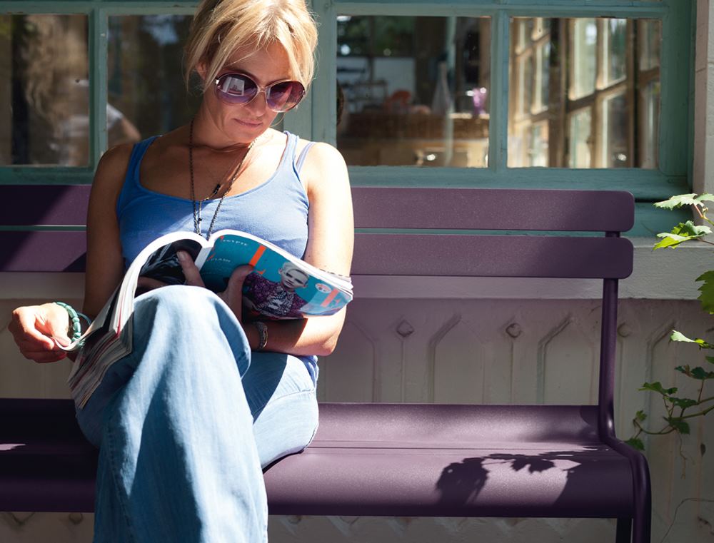 Frau liest auf lila Luxembourg Bank von Fermob im Freien ein Magazin.