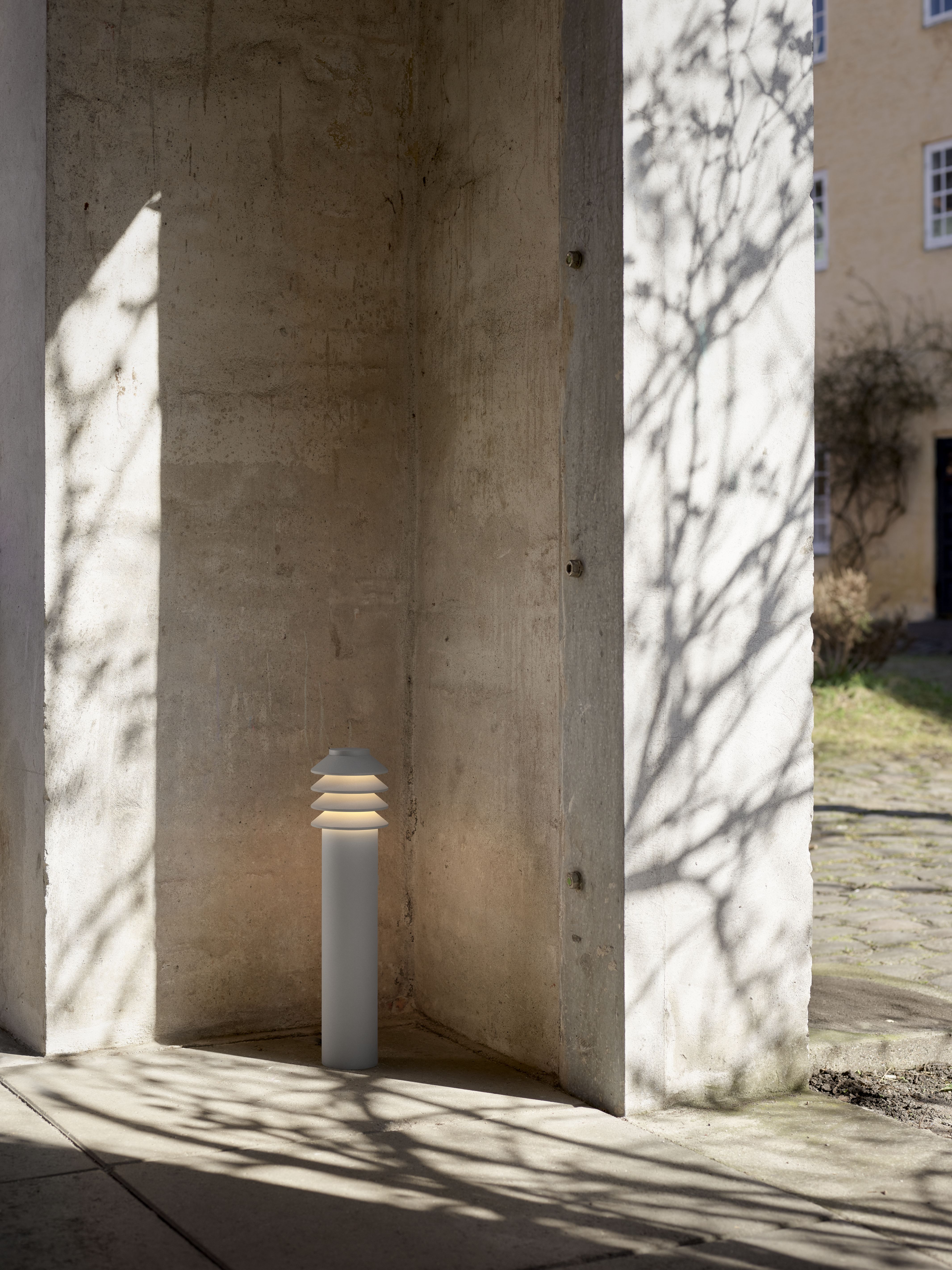 Bysted Garden Pollerleuchte von Louis Poulsen im Außenbereich, moderne Gartenbeleuchtung mit Baumschatten auf Beton.