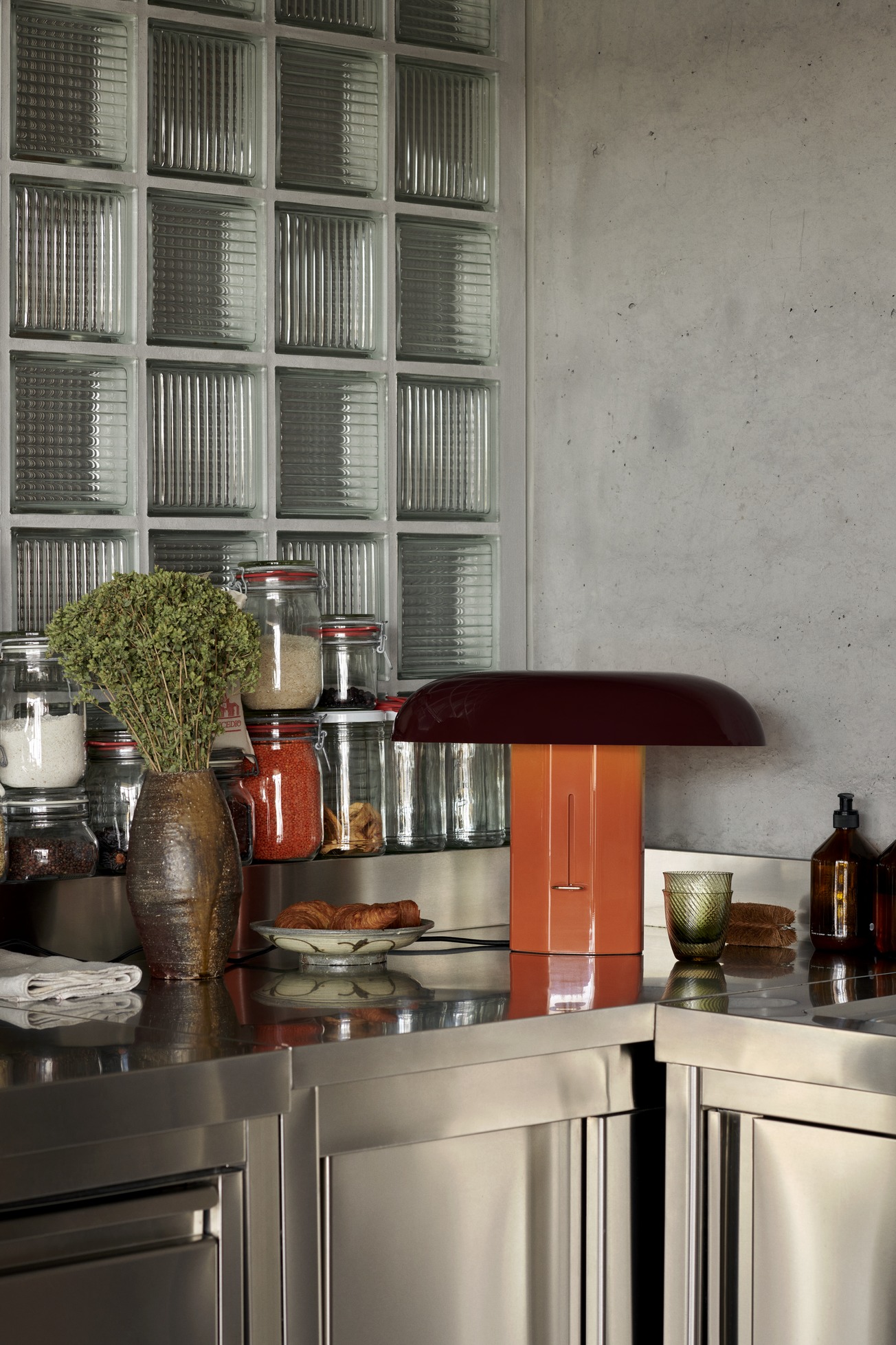 AndTradition Montera JH42 Tischleuchte in Orange und Braun auf einer Küchentheke mit Glasbausteinen im Hintergrund.