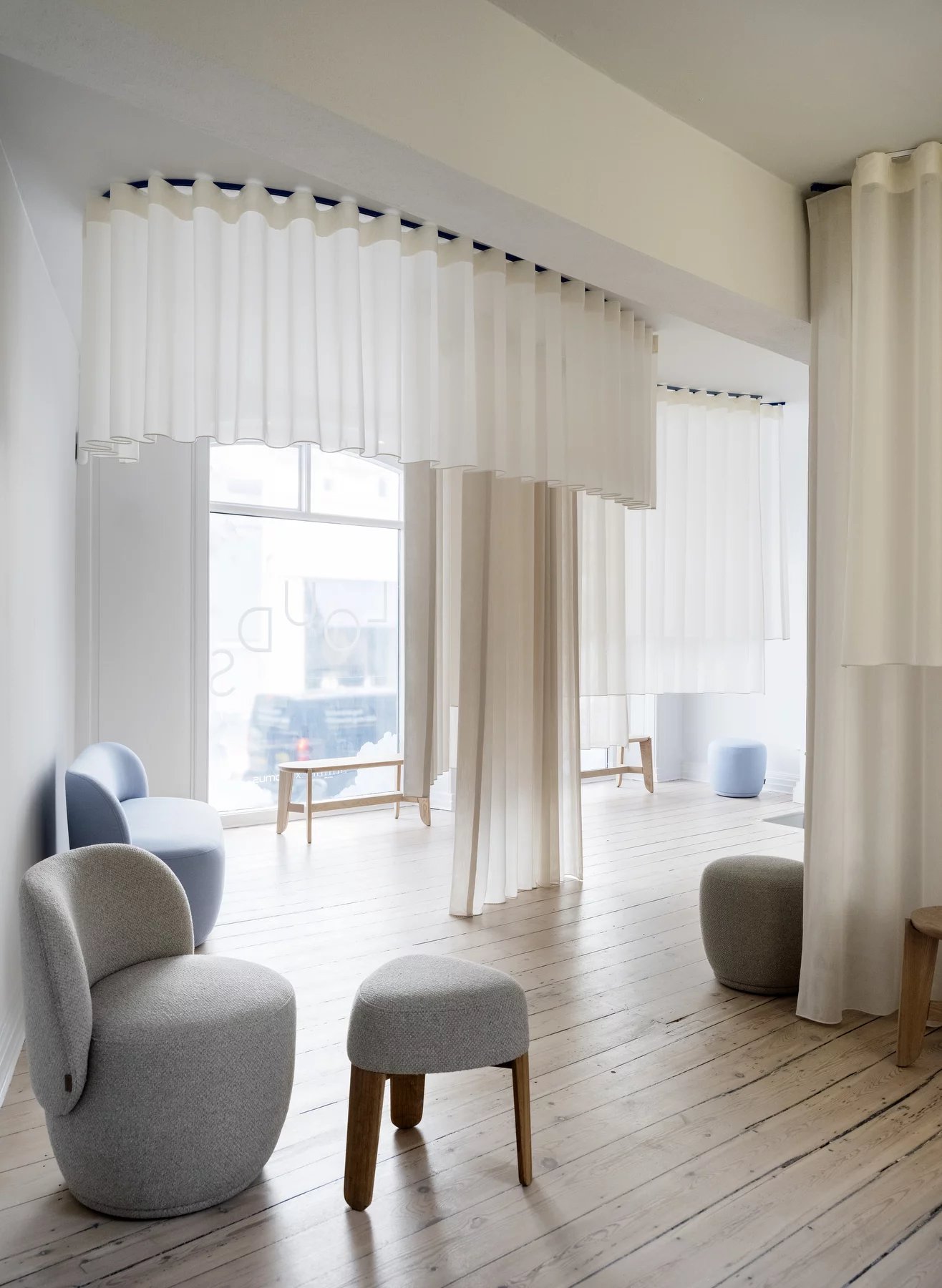 Innenaufnahme mit Blomus Socia Sesseln und Hockern in Taupe und Blau vor weißen Vorhängen.