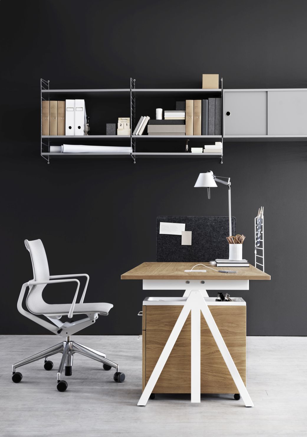 Works Schreibtisch höhenverstellbar B140cm von String Furniture mit Regalsystem, Bürostuhl und Schreibtischlampe vor dunkler Wand.