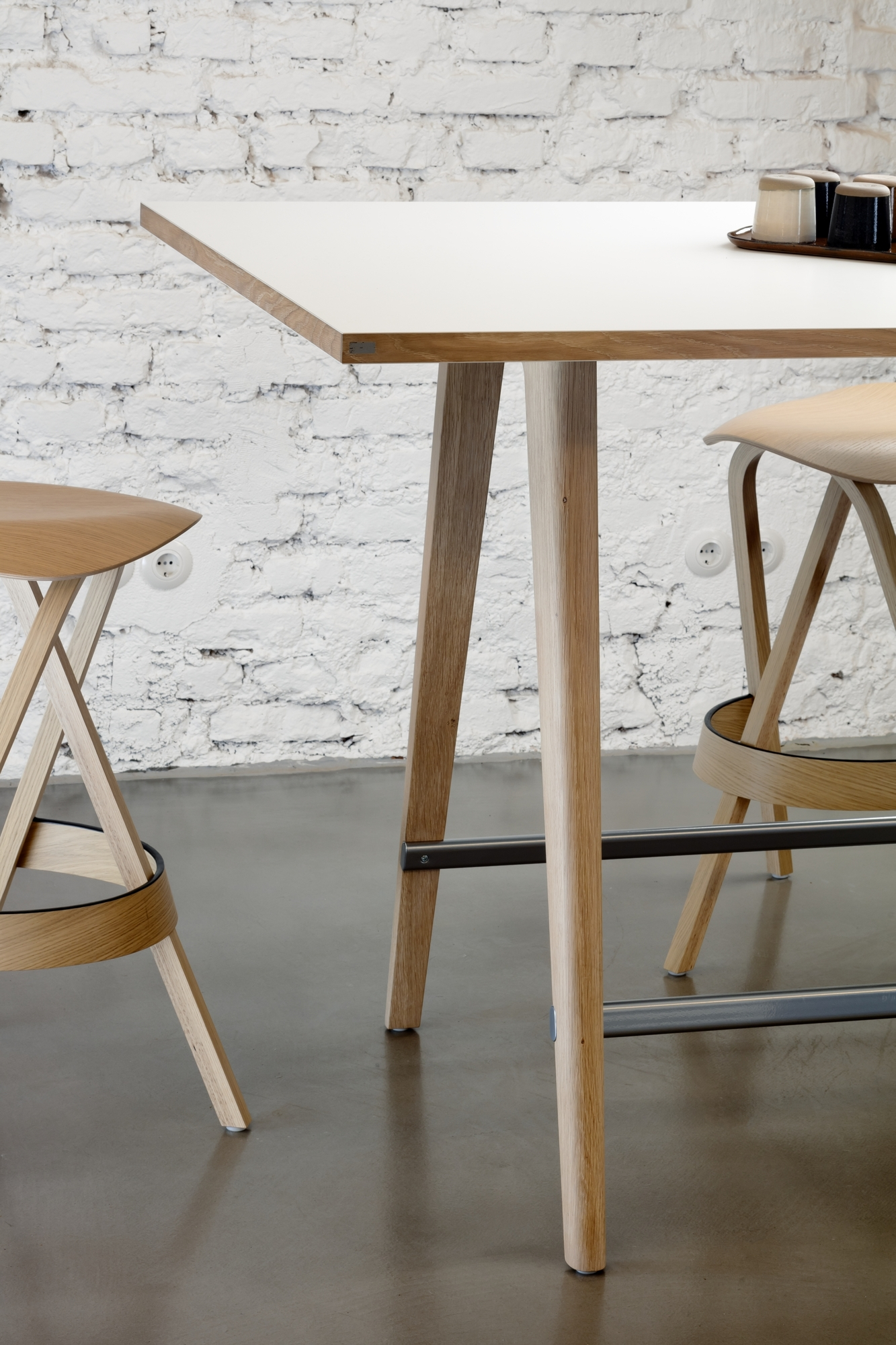 Detailaufnahme des Thonet 1510 Stehtisches mit Barhockern, hellem Holzgestell und weißer Tischplatte vor einer Backsteinwand.