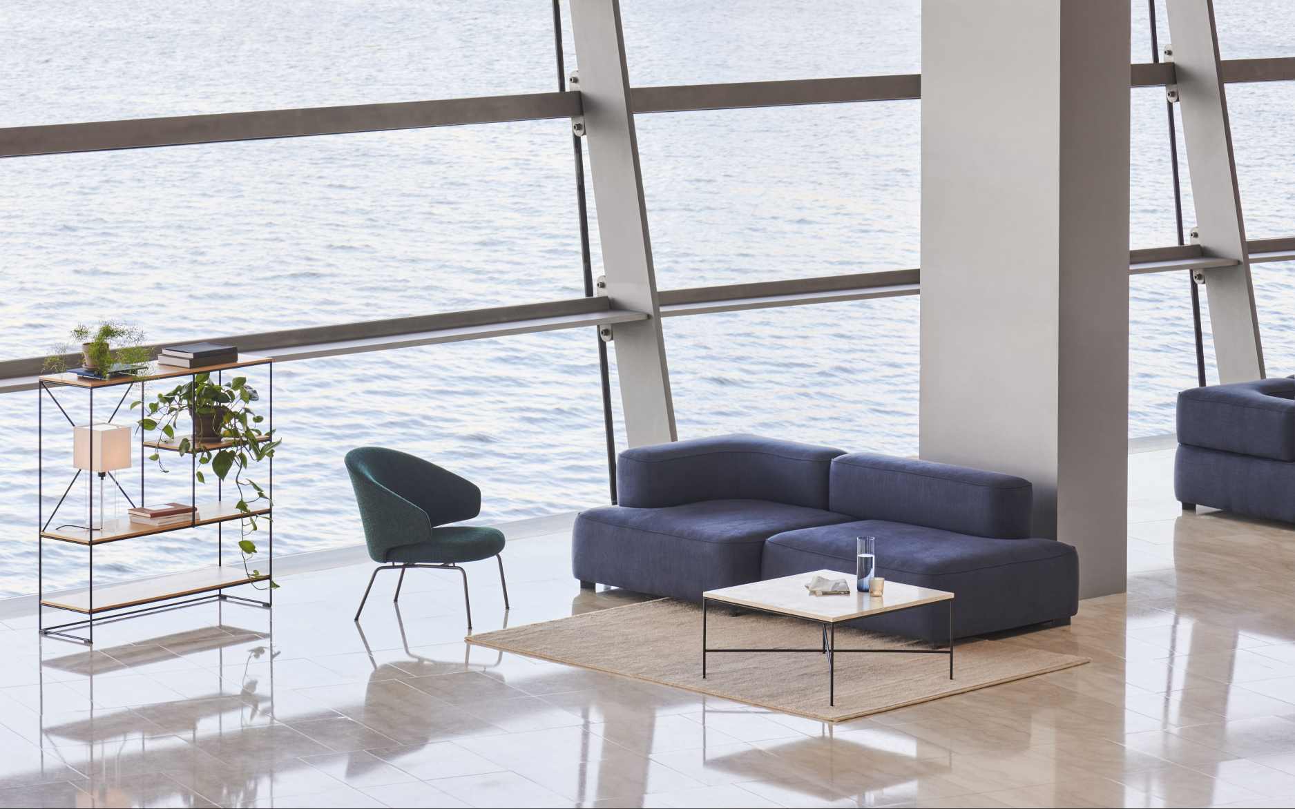 Moderner Wohnraum mit blauem Ecksofa, Couchtisch und grünem Fritz Hansen Let Sessel vor Fensterfront mit Meerblick.