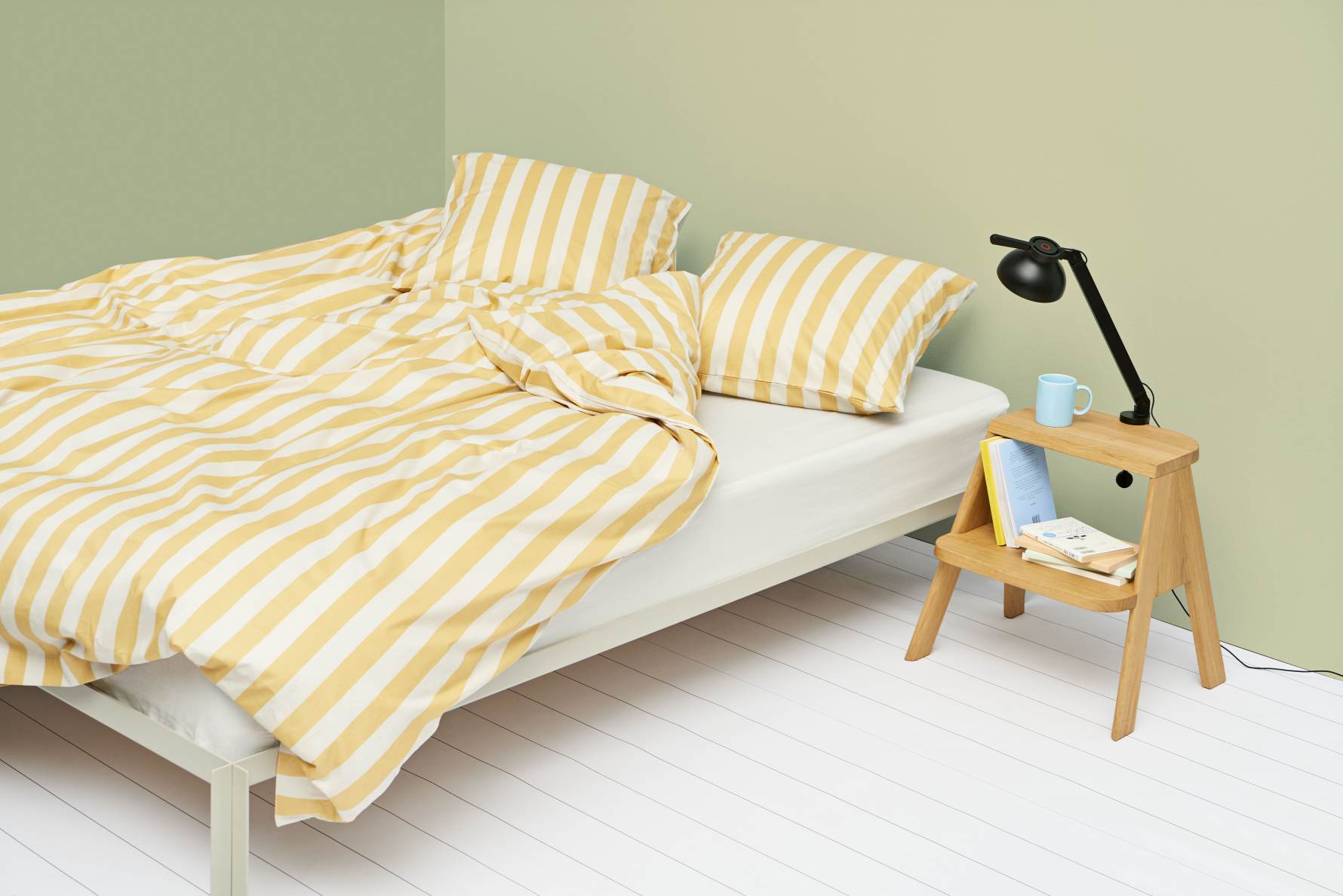 Heller Raum mit Bett und gelb-weiß gestreifter Bettwäsche, daneben ein Hay Butler Hocker als Nachttisch.
