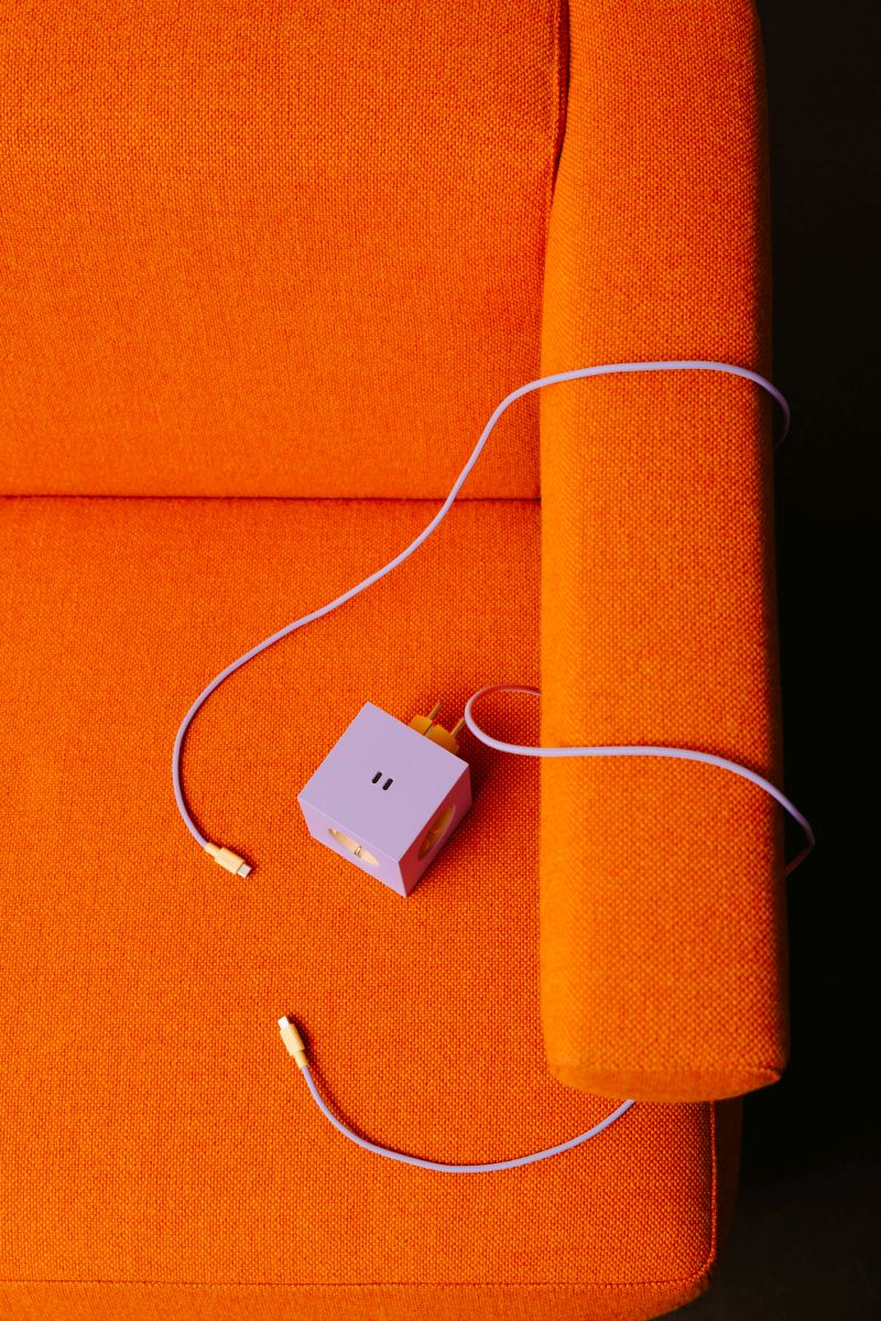 Lila Avolt Square 2 Steckdosenwürfel auf einem orangefarbenen Sessel mit USB-C Kabel.