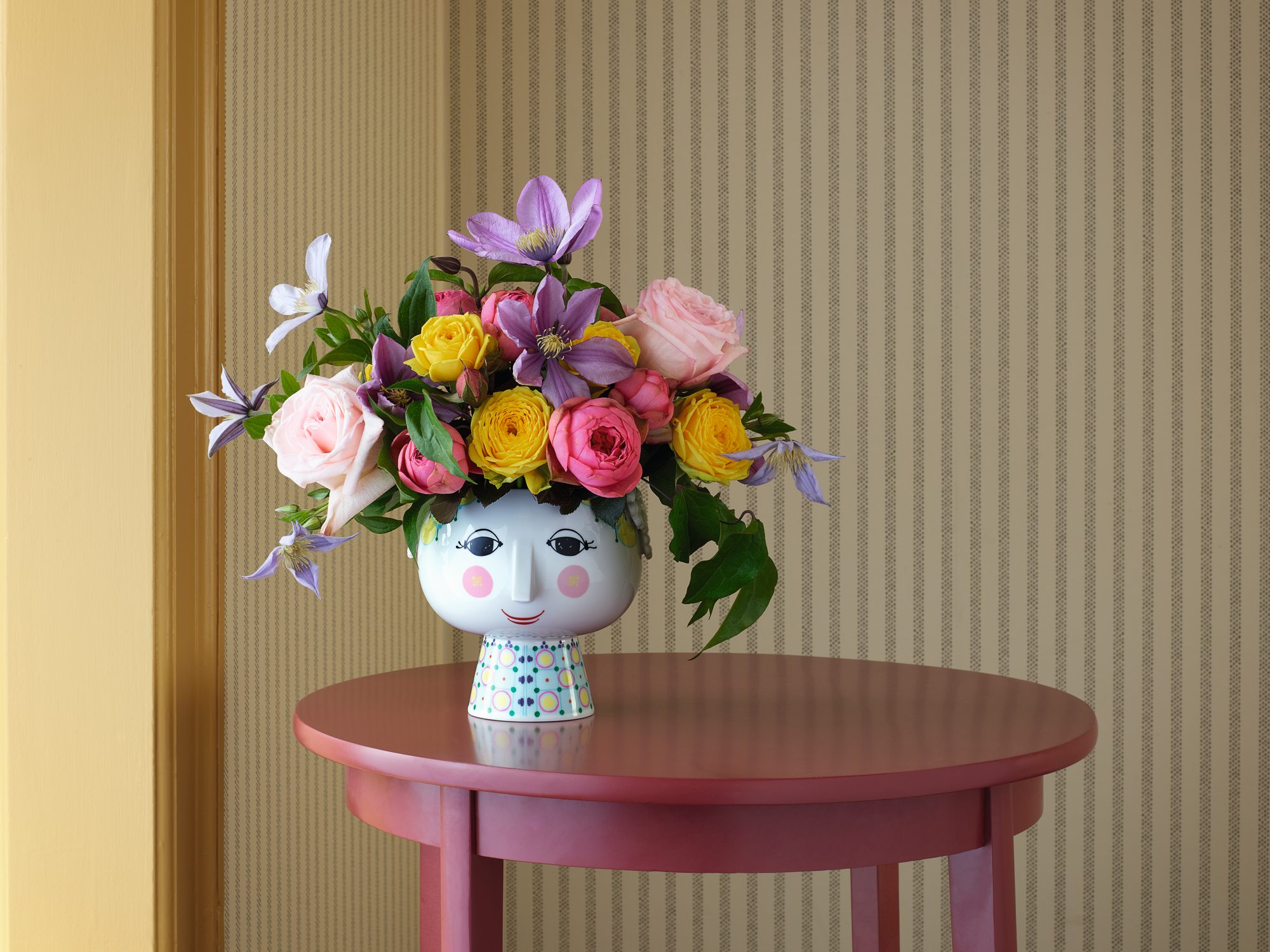 Übertopf Eva Bjorn Wiinblad mit bunten Blumen auf einem runden, roten Tisch. Keramik Übertopf im Gesicht Design.