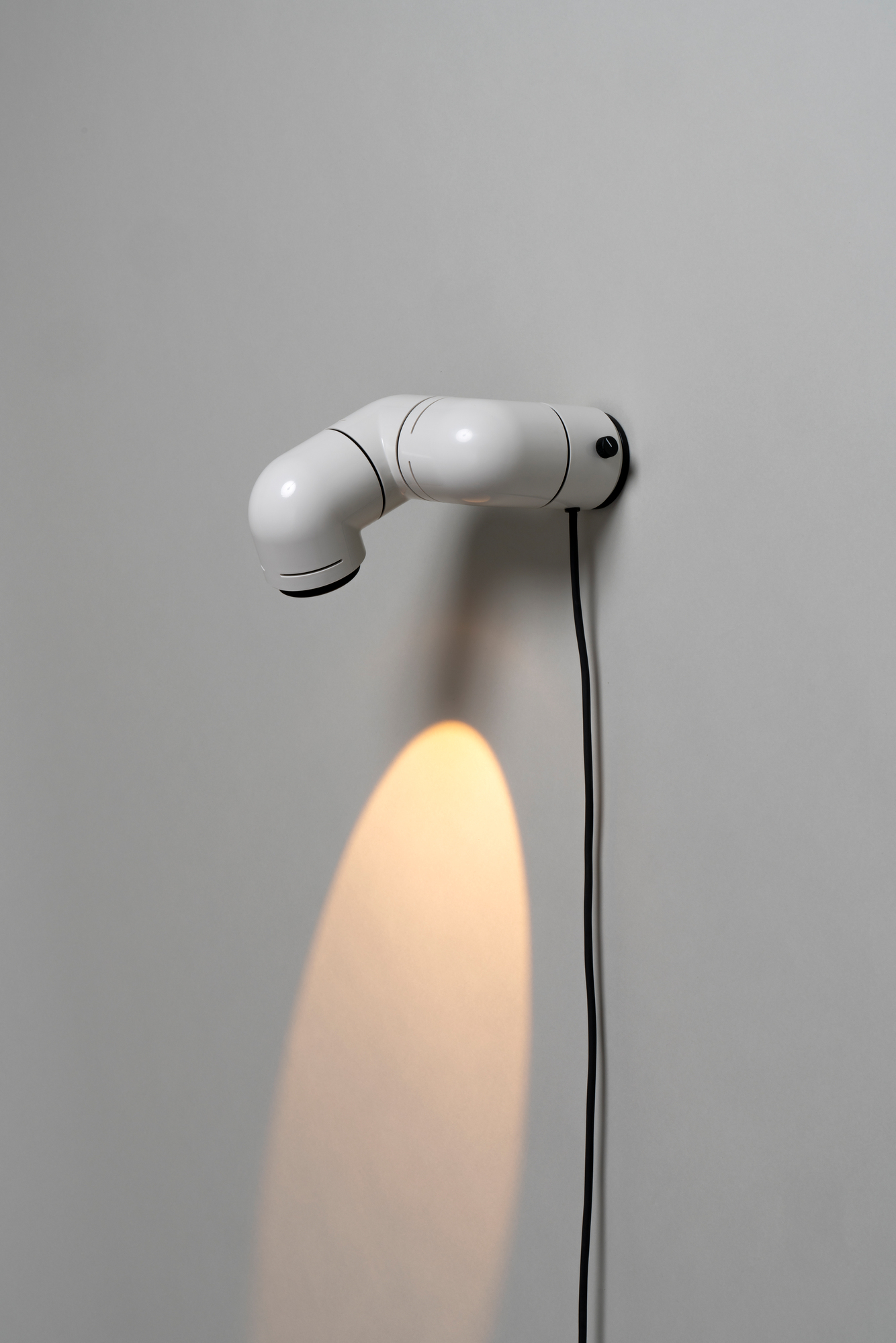 Weiße Tatu Tischleuchte von Santa & Cole an einer Wand mit Lichtkegel und schwarzem Kabel.