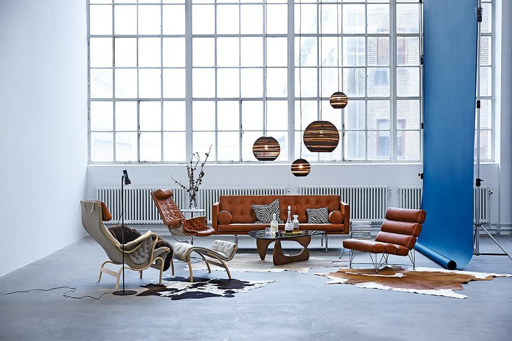 Wohnzimmer mit Graypants Moon18 Scraplights Hängeleuchten, braunem Ledersofa und Sesseln vor großem Fenster.