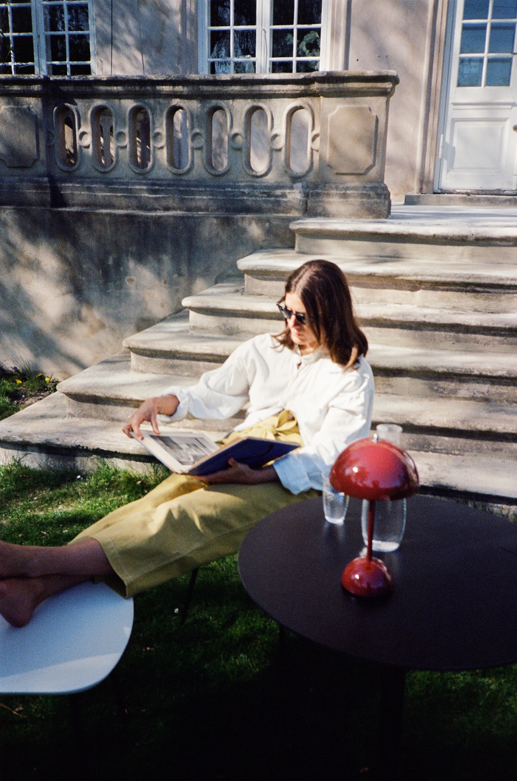 Frau liest im Garten auf dem AndTradition Rely HW70 Outdoor Chair Stuhl mit Tisch und Lampe.