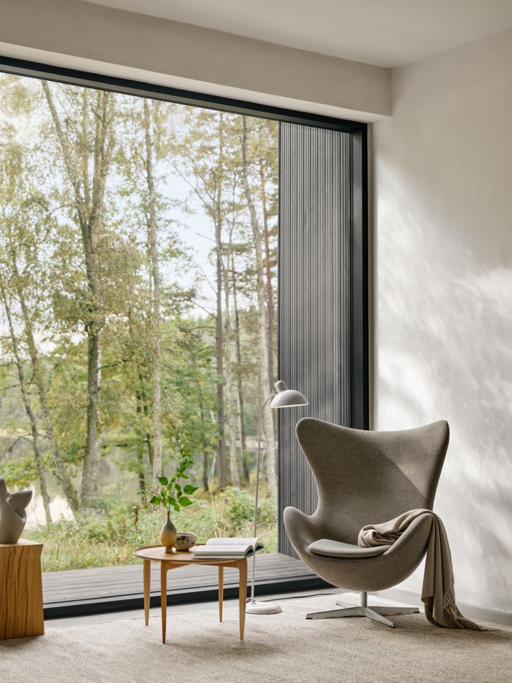 Fritz Hansen Tray Beistelltisch Ø60cm aus Holz neben Sessel und Stehlampe vor Fenster mit Waldblick.