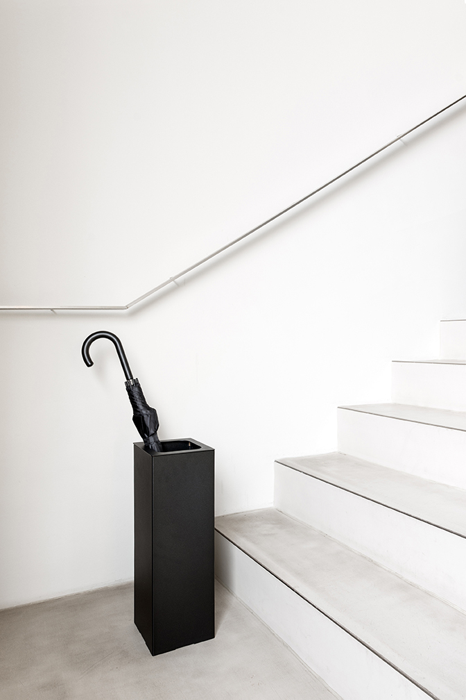 Schwarzer Angle Schirmständer von Schönbuch mit Regenschirm neben einer Treppe in minimalistischem Design.