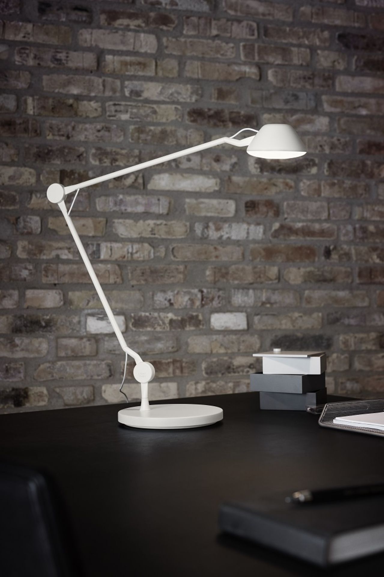 Weiße AQ01 Tischleuchte von Fritz Hansen auf einem Schreibtisch vor einer Backsteinwand.