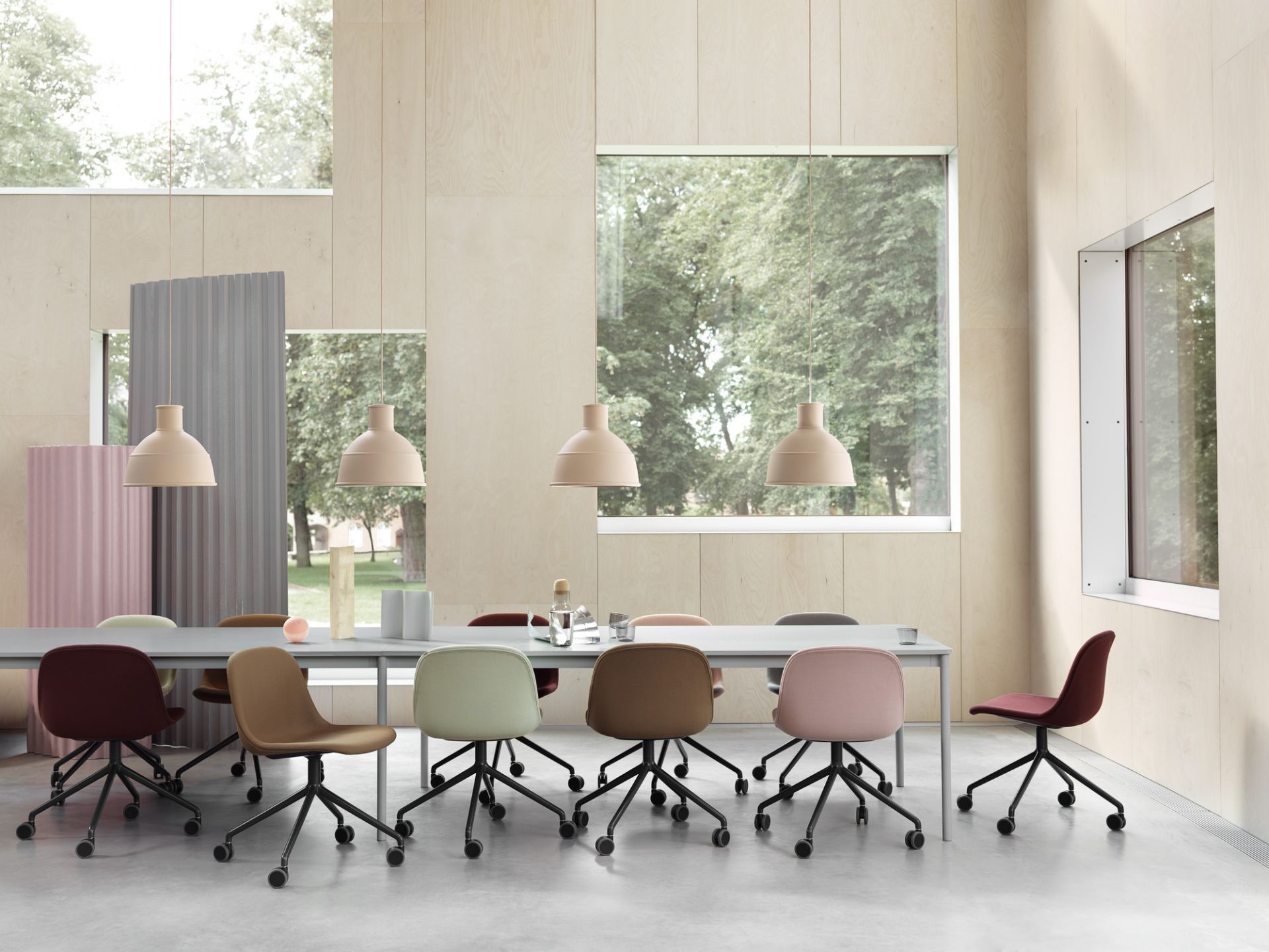 Helles Büro mit langem Tisch und farbigen Stühlen, darüber hängen Lampen. Muuto Tisch und Stühle.