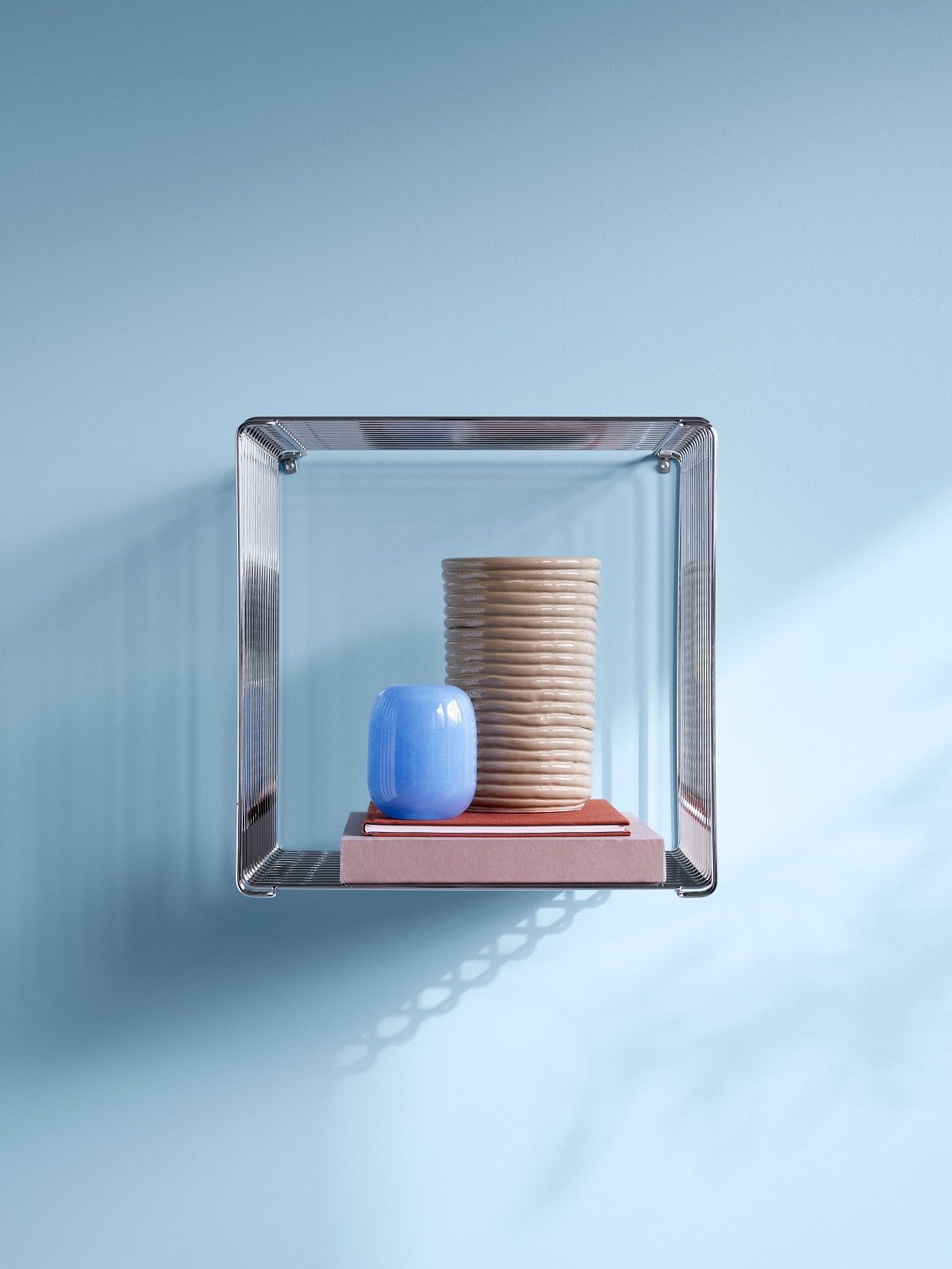 Montana Panton Wire Cube Single Drahtregal in Chrom mit Deko-Objekten vor hellblauer Wand.