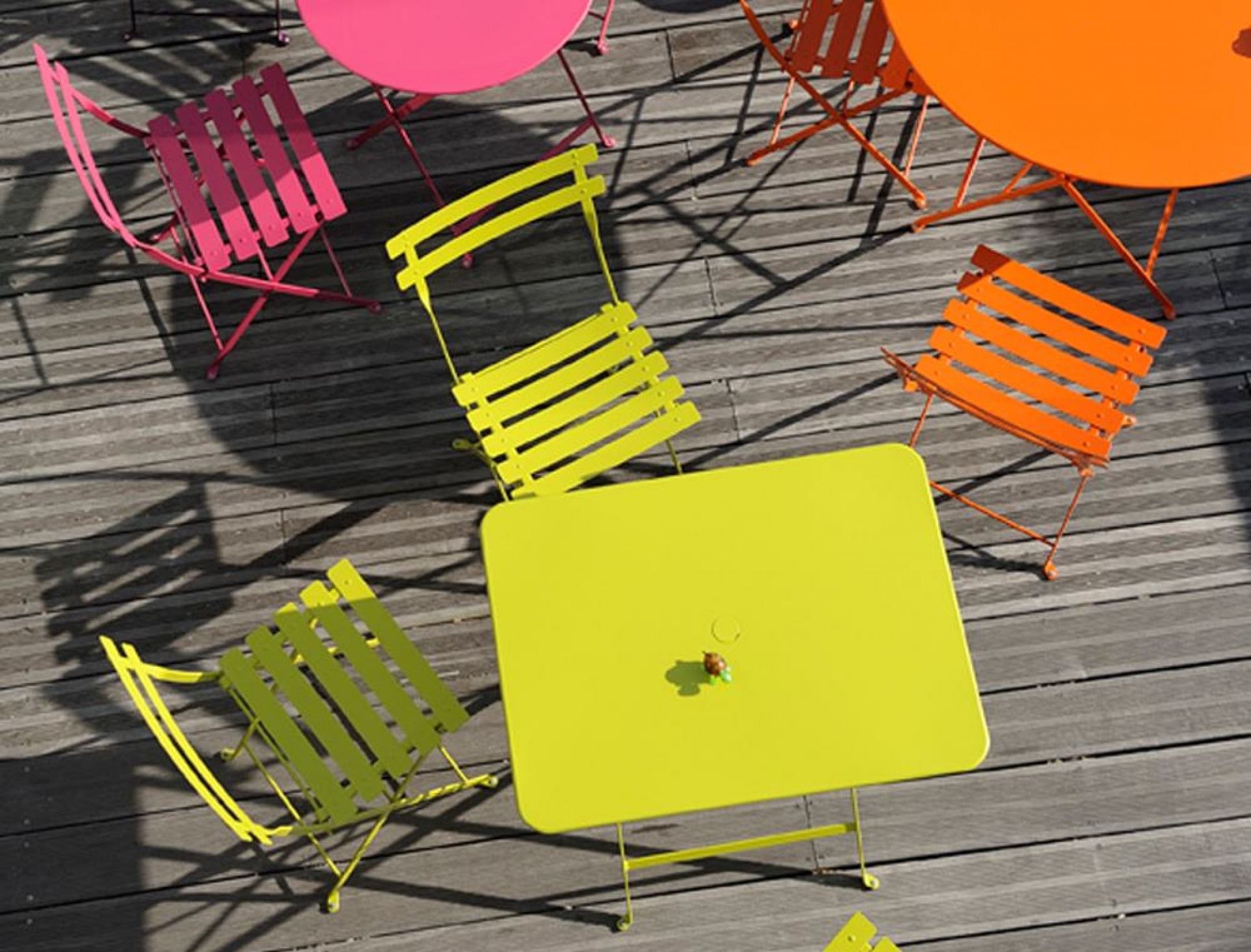 Draufsicht auf farbenfrohe Fermob Bistro Gartenmöbel: Klapptische und Stühle in Pink, Gelb und Orange auf Holzterrasse.