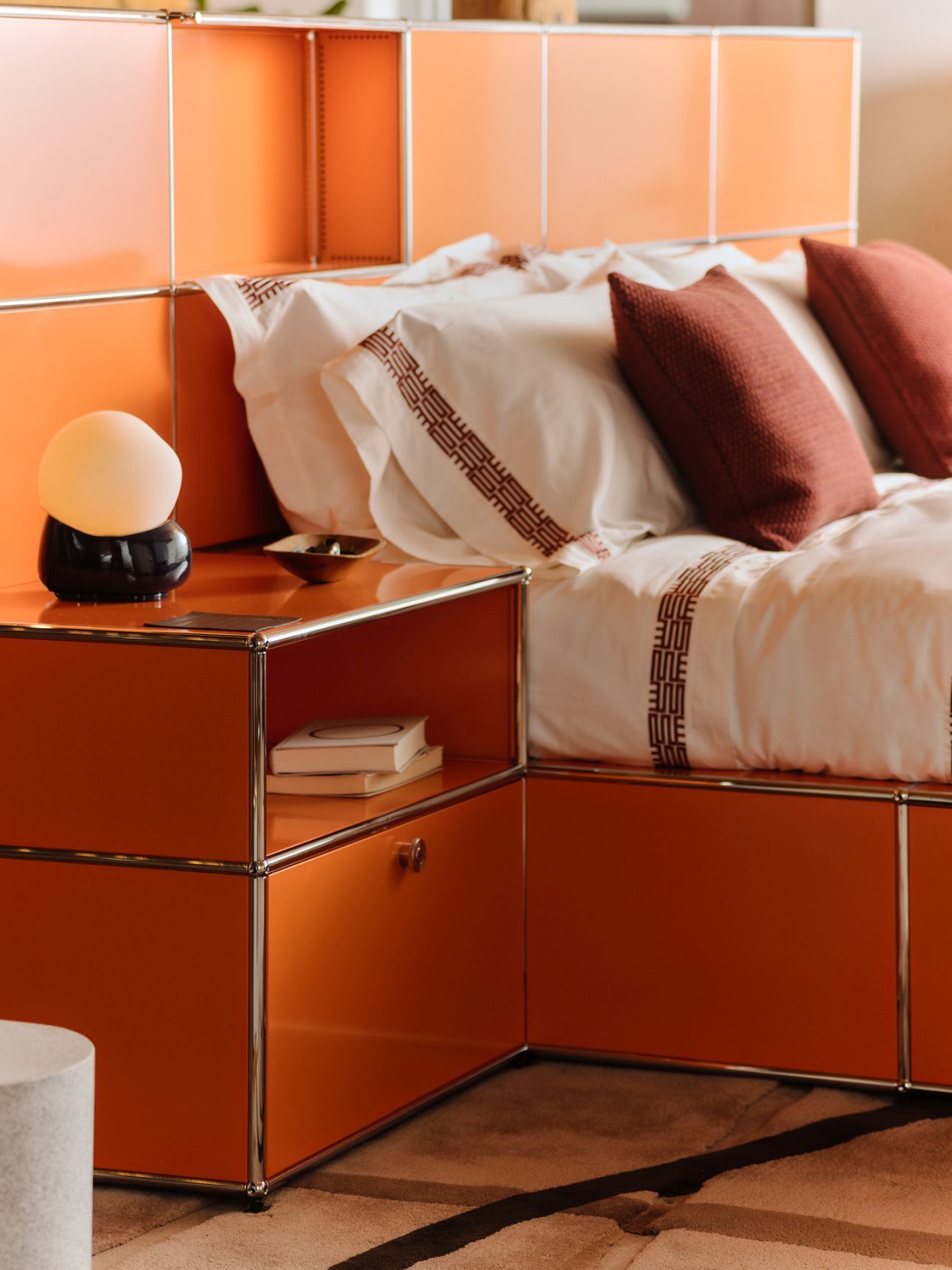 Orangefarbenes USM Haller Bett Mansa mit Nachttisch, Lampe, Kissen und weißer Bettwäsche im Schlafzimmer.