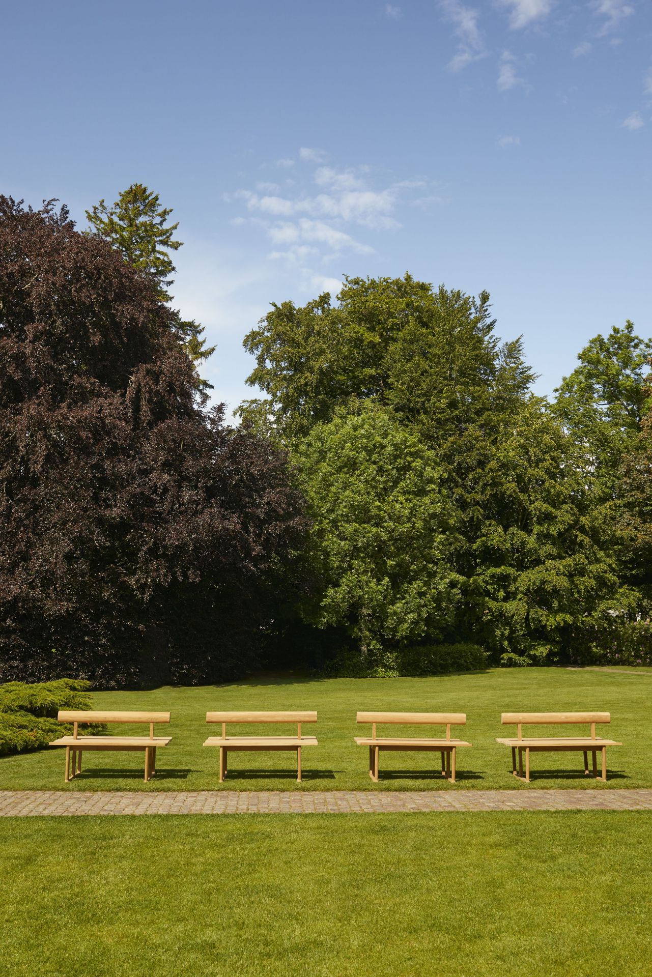 Vier Skagerak Banco Teak Gartenbänke auf Rasen vor Bäumen unter blauem Himmel, Outdoor Möbel.