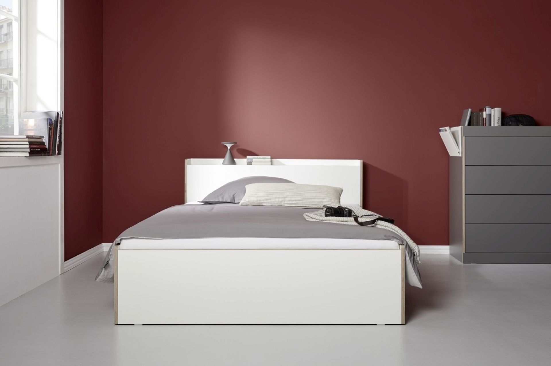 Nook Einzelbett von Müller Möbelwerkstätten in Weiß mit grauem Bettzeug vor bordeauxroter Wand.