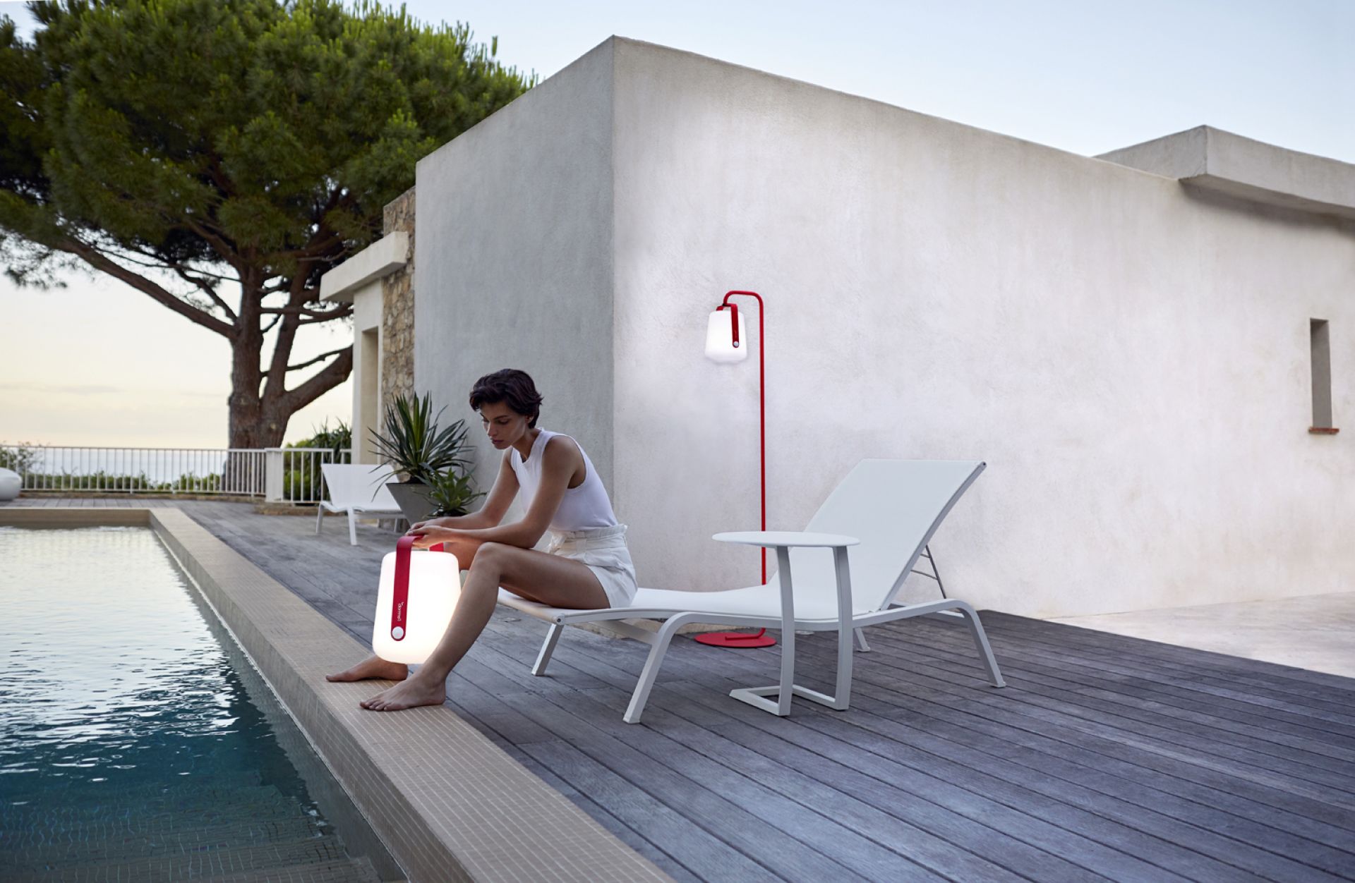 Frau sitzt am Pool mit Alizé Outdoor Tischleuchte und roter Stehlampe von Fermob im modernen Garten.