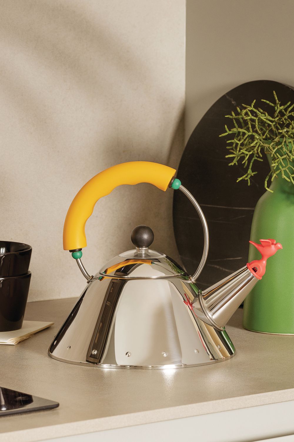 Alessi Wasserkessel 9093/1 Y mit gelbem Griff und Vogelpfeife aus Edelstahl auf Küchentisch.