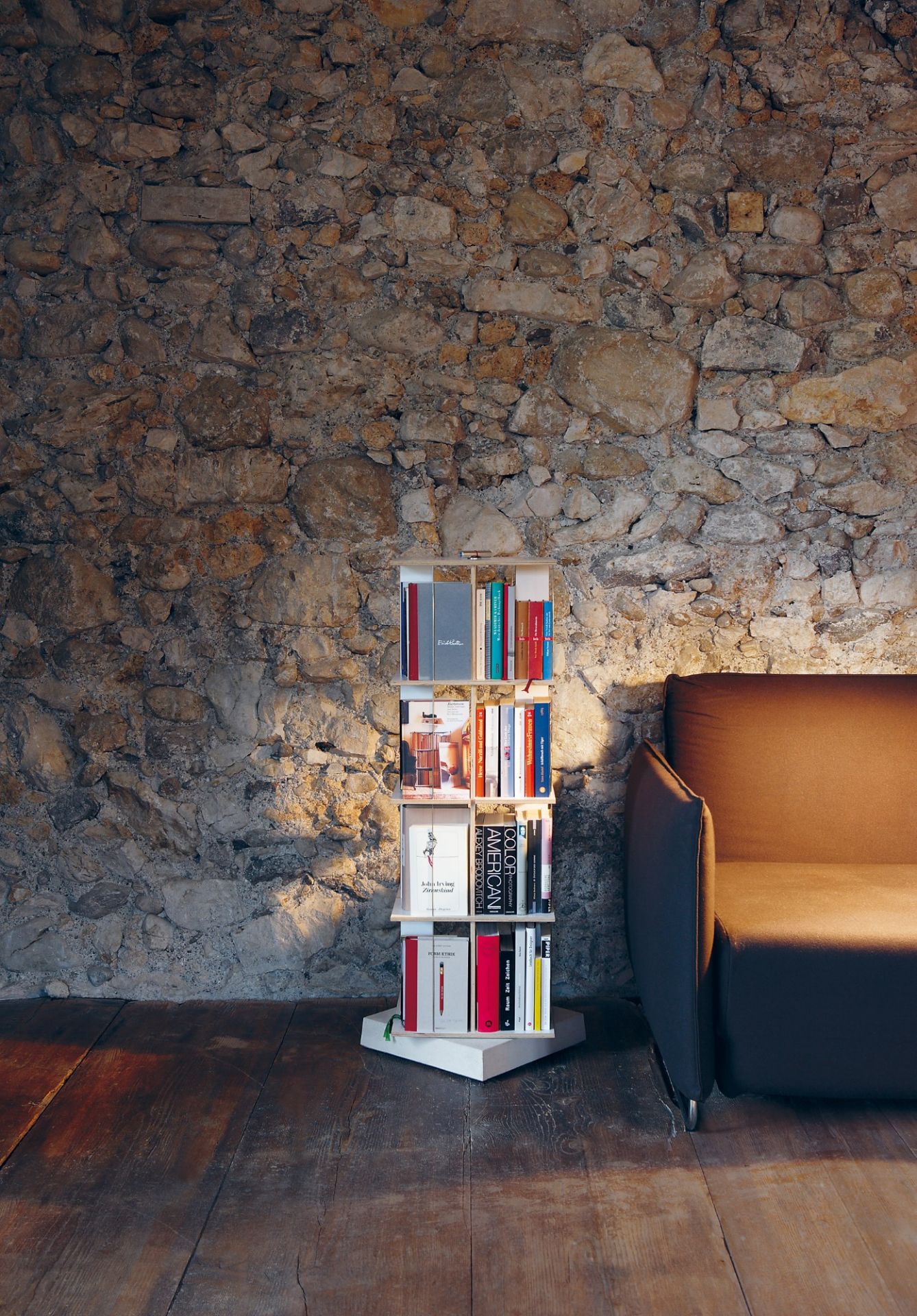 Nils Holger Moormann Bücherregal: Der Buchstabler Bücherturm vor einer Steinwand und einem Sofa.