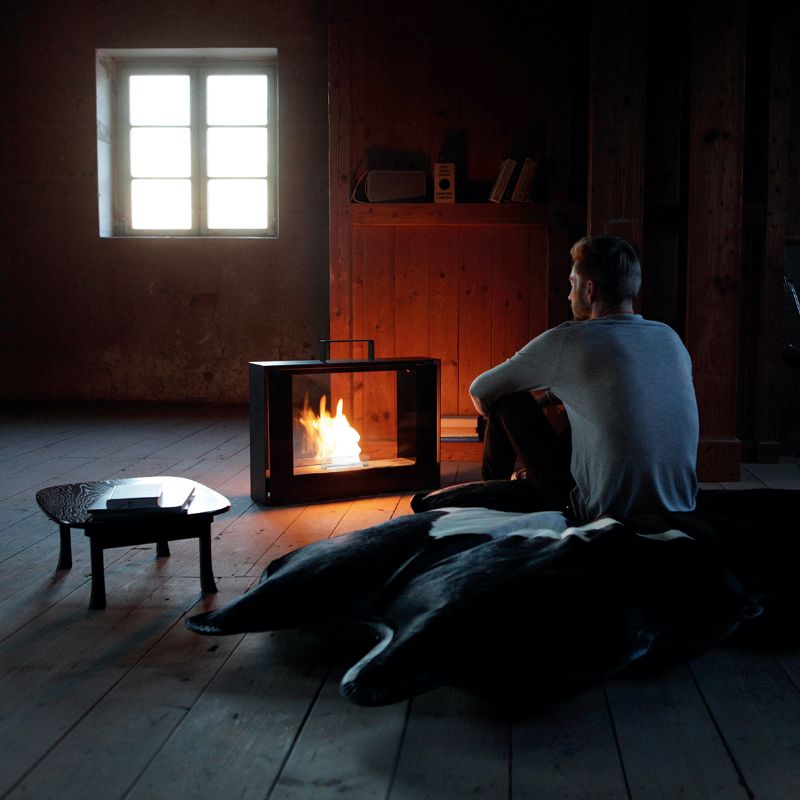 Travelmate Conmoto Kamin mit Tenderflame Brenner im dunklen Raum, Mann sitzt daneben und genießt die Wärme.