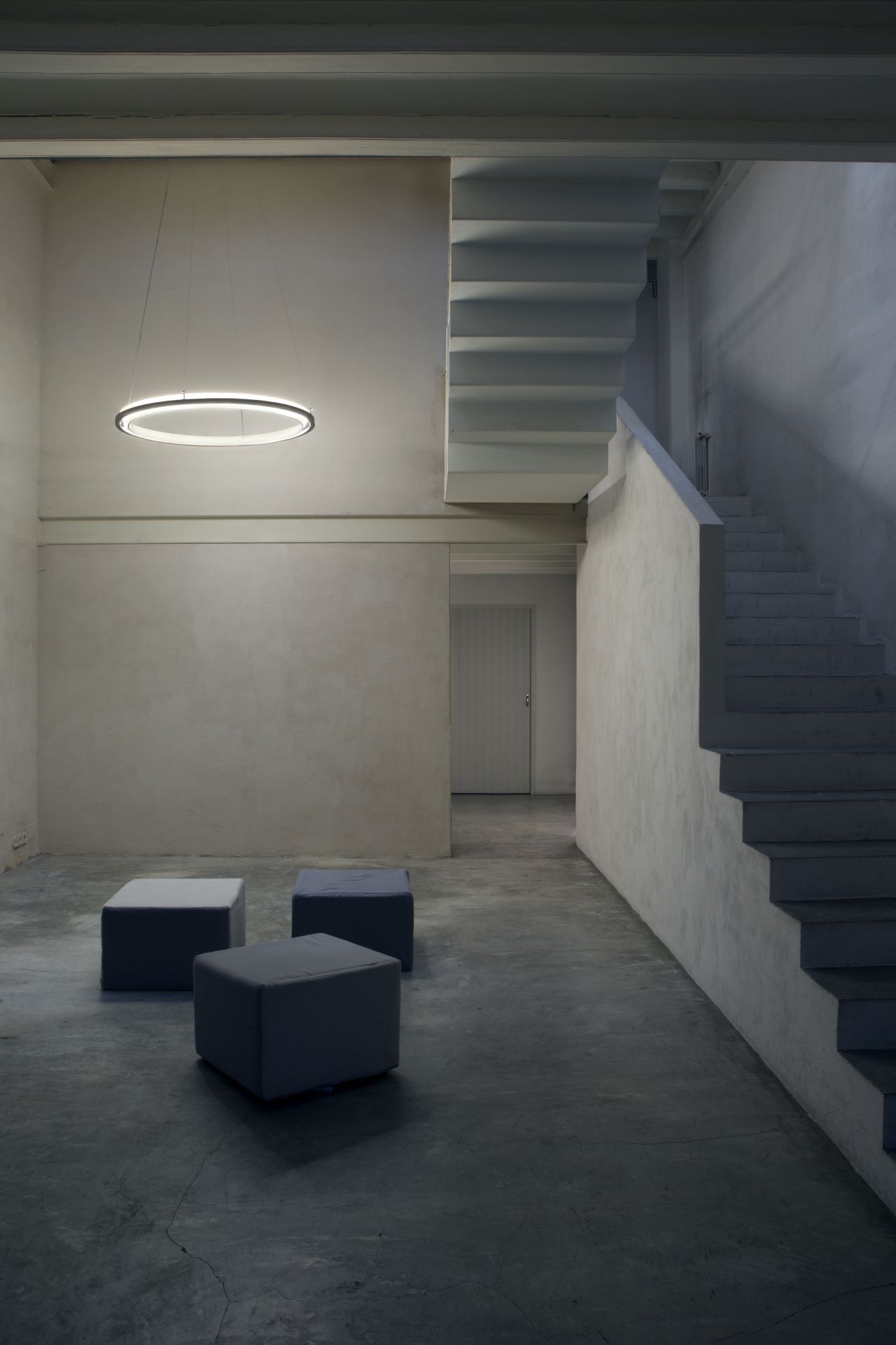 Moderne Ring-Hängeleuchte über grauen Sitzhockern in minimalistischem Raum mit Treppe und Betonboden.