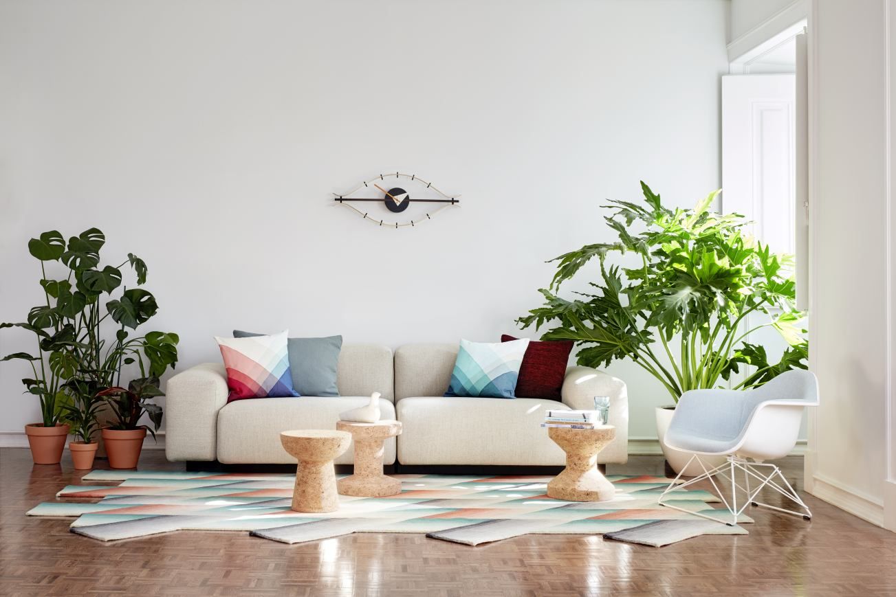 Helle Wohnzimmer-Szene mit Vitra Eye Clock Wanduhr über einem beigen Sofa und Pflanzen.