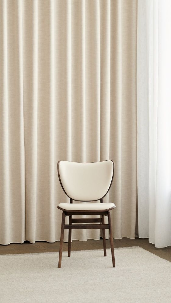 Vollgepolsterter Elephant Dining Chair Stuhl von NORR11 mit Holzrahmen und beigem Bezug vor Vorhang.