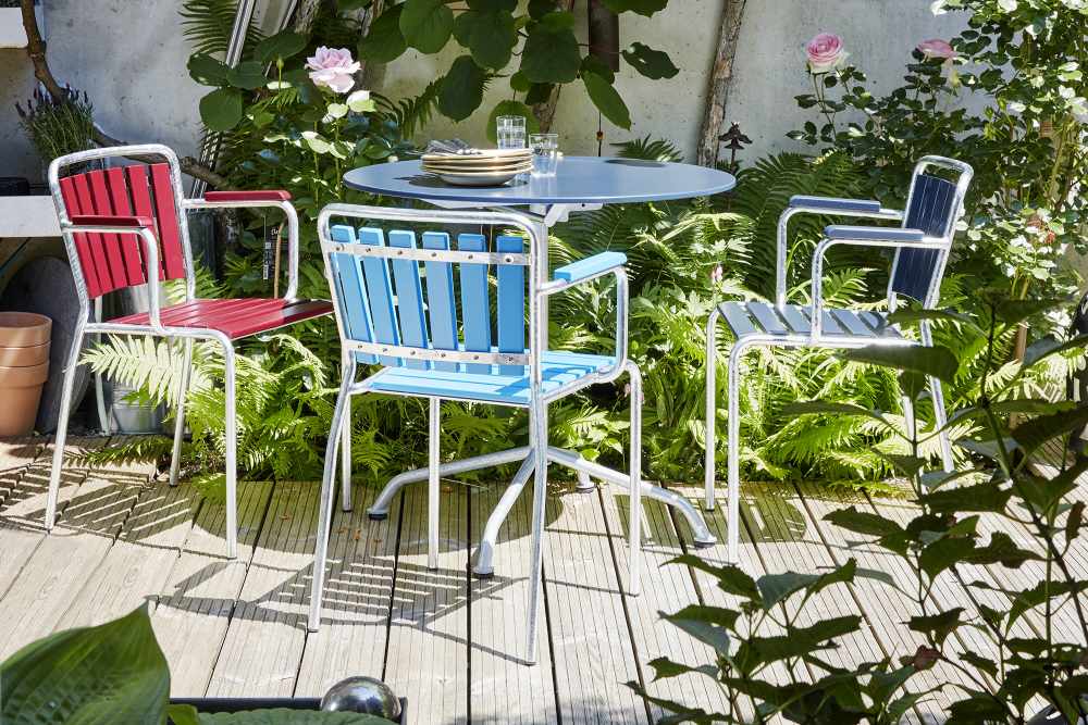 Gelber Haefeli Gartentisch mit Stühlen auf einer Holzterrasse, umgeben von üppigem Grün. Outdoor-Möbel für Garten und Balkon.
