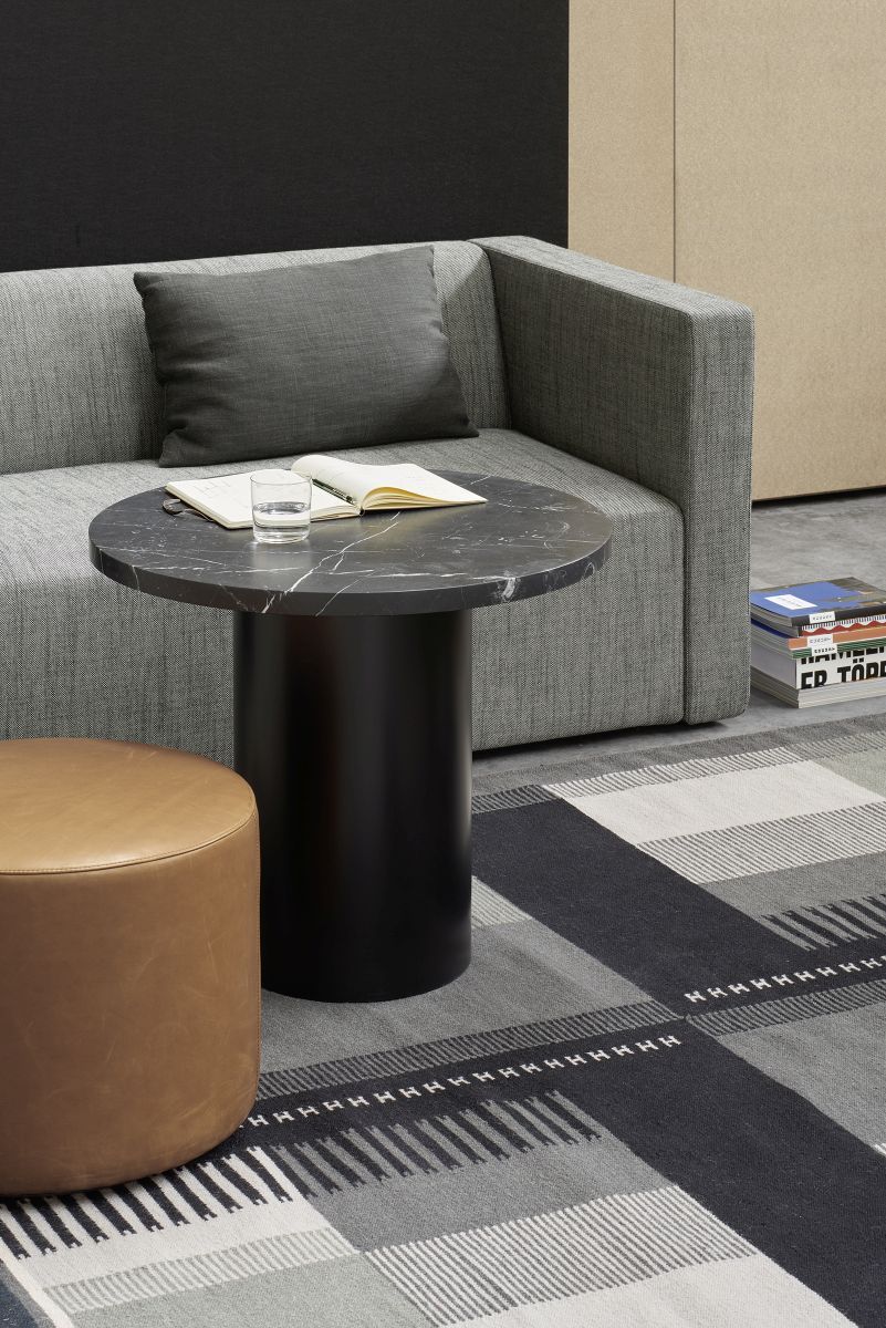 Enoki CT09 Beistelltisch von e15 mit Marmorplatte und schwarzem Zylinderfuß neben grauem Sofa und Lederhocker.