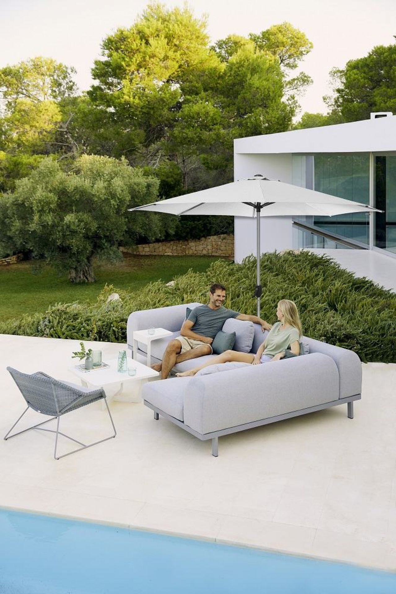 Paar entspannt unter Cane-Line Sunshade Parasol Sonnenschirm mit Kurbel auf grauer Gartenlounge am Pool.
