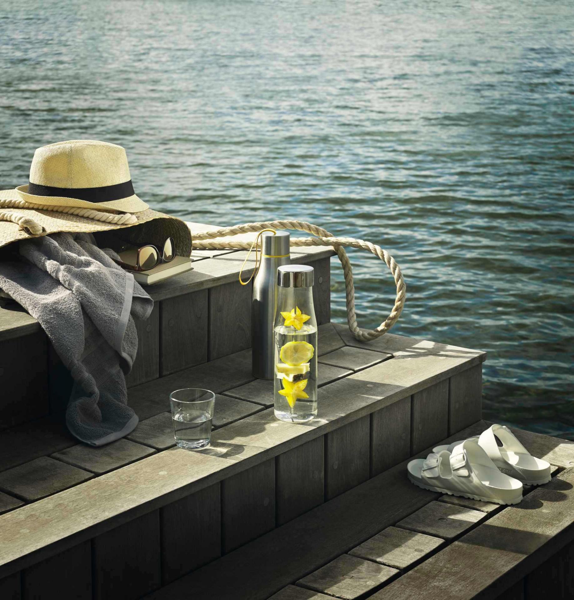 My Flavour Karaffe von Eva Solo auf Holzsteg mit Wasser, Zitrone und Sternfrucht, neben Strandutensilien.