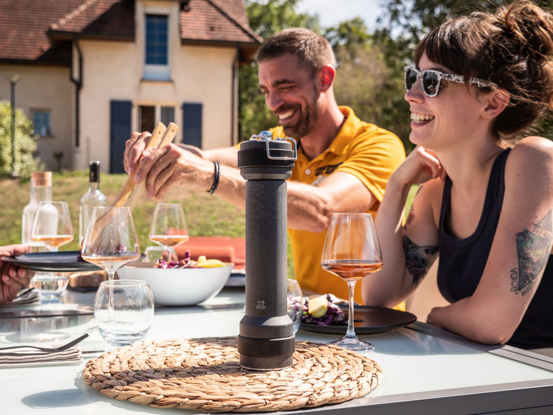 Graphitfarbene Peugeot Pfeffermühle auf einem Tisch mit lachenden Menschen im Hintergrund für ein BBQ.