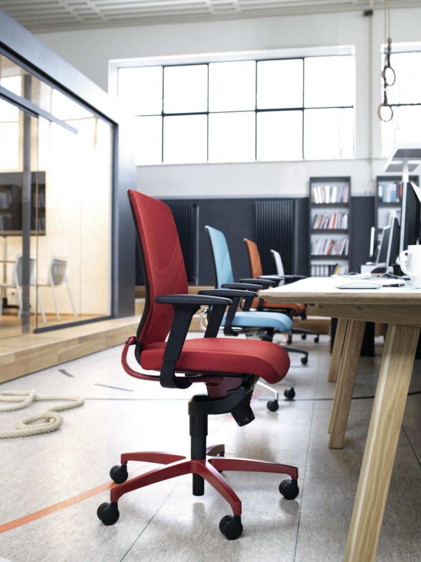 Roter Bürostuhl mit Armlehnen und Rollen im modernen Büro, weitere Bürostühle in blau und braun.