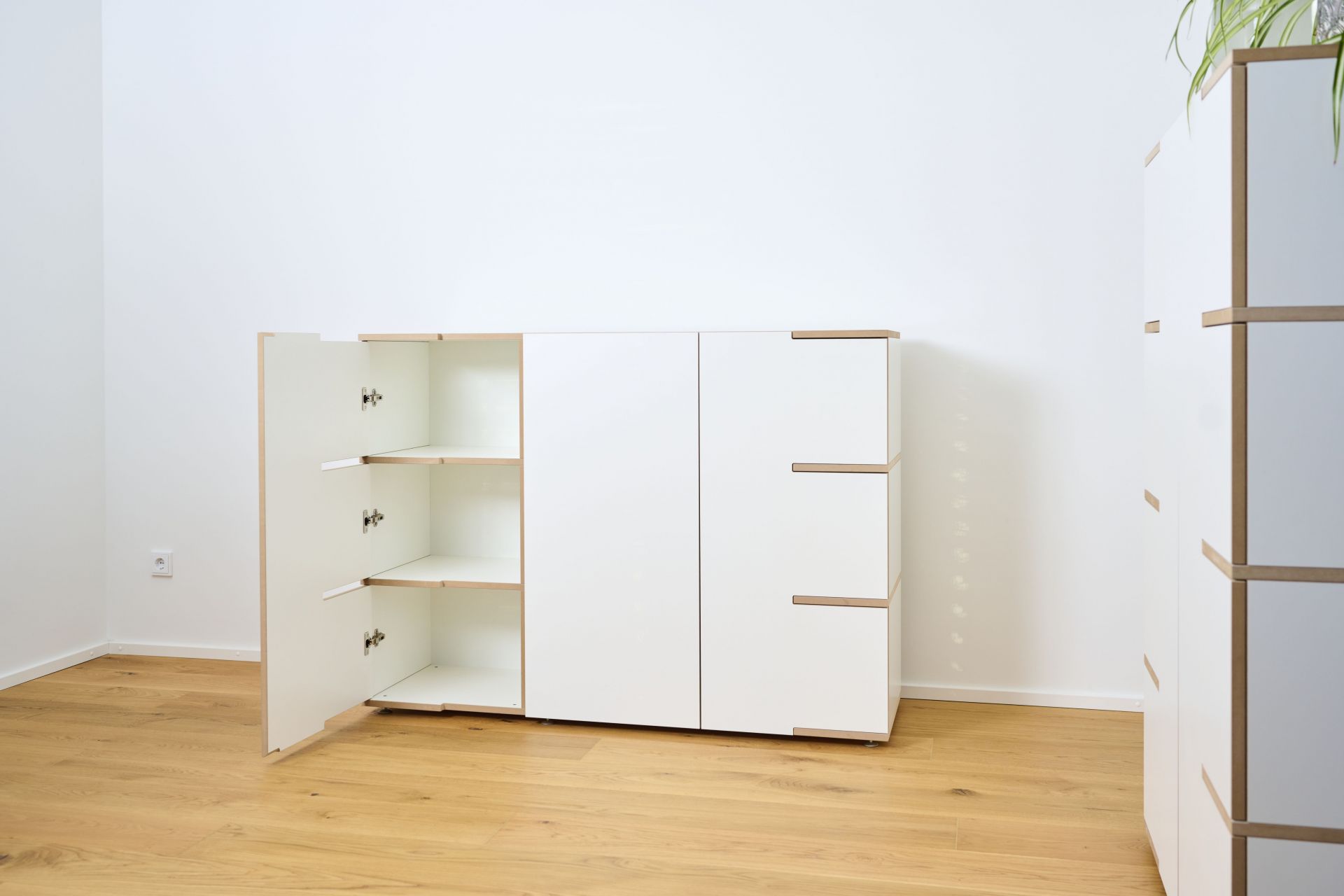 Weißes Tojo Stau Sideboard mit offener Tür und drei Regalböden auf hellem Holzboden.